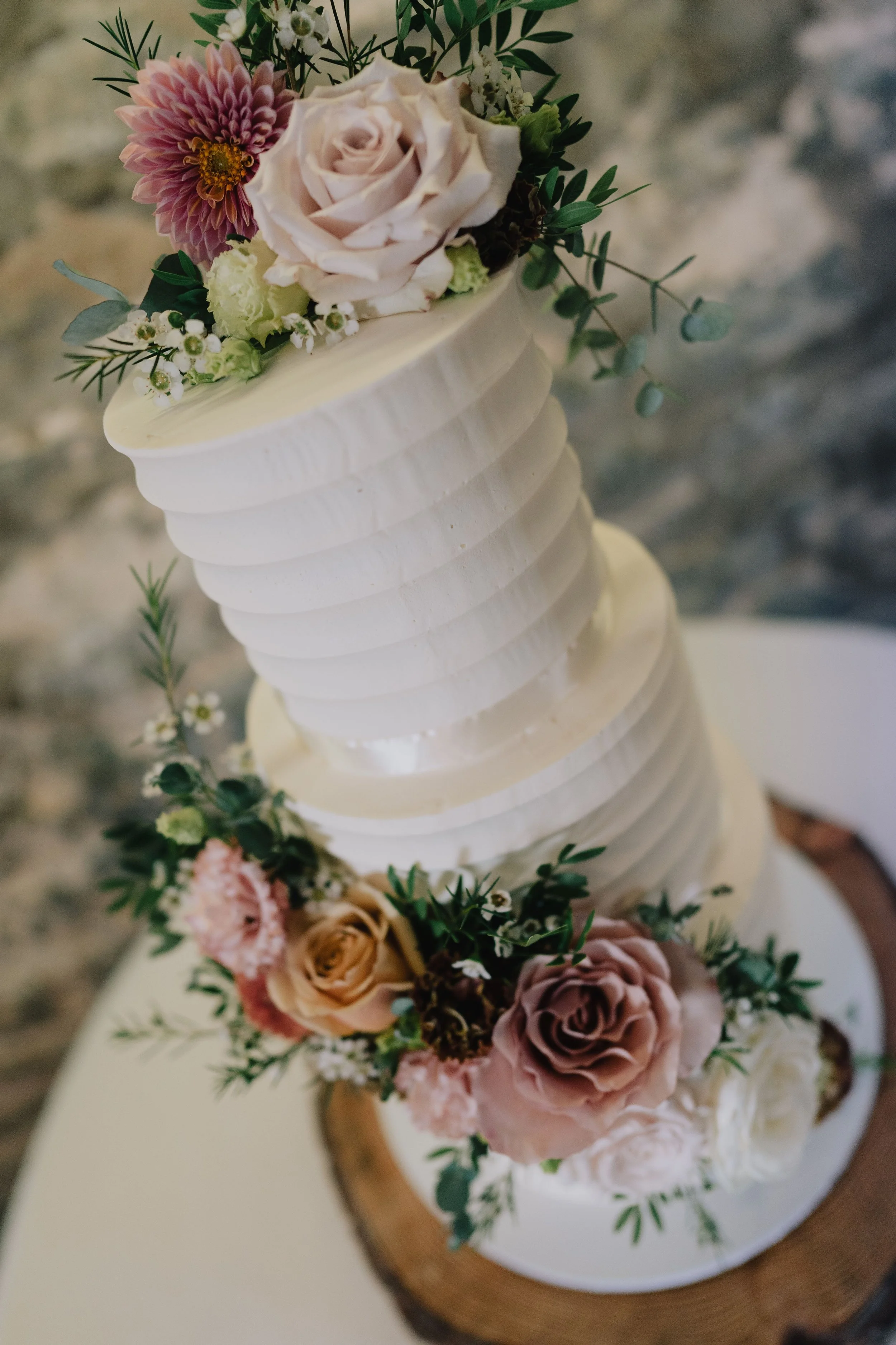 A tall, elegant white wedding cake decorated with fresh pink roses, white roses, and greenery, placed on a wooden stand.