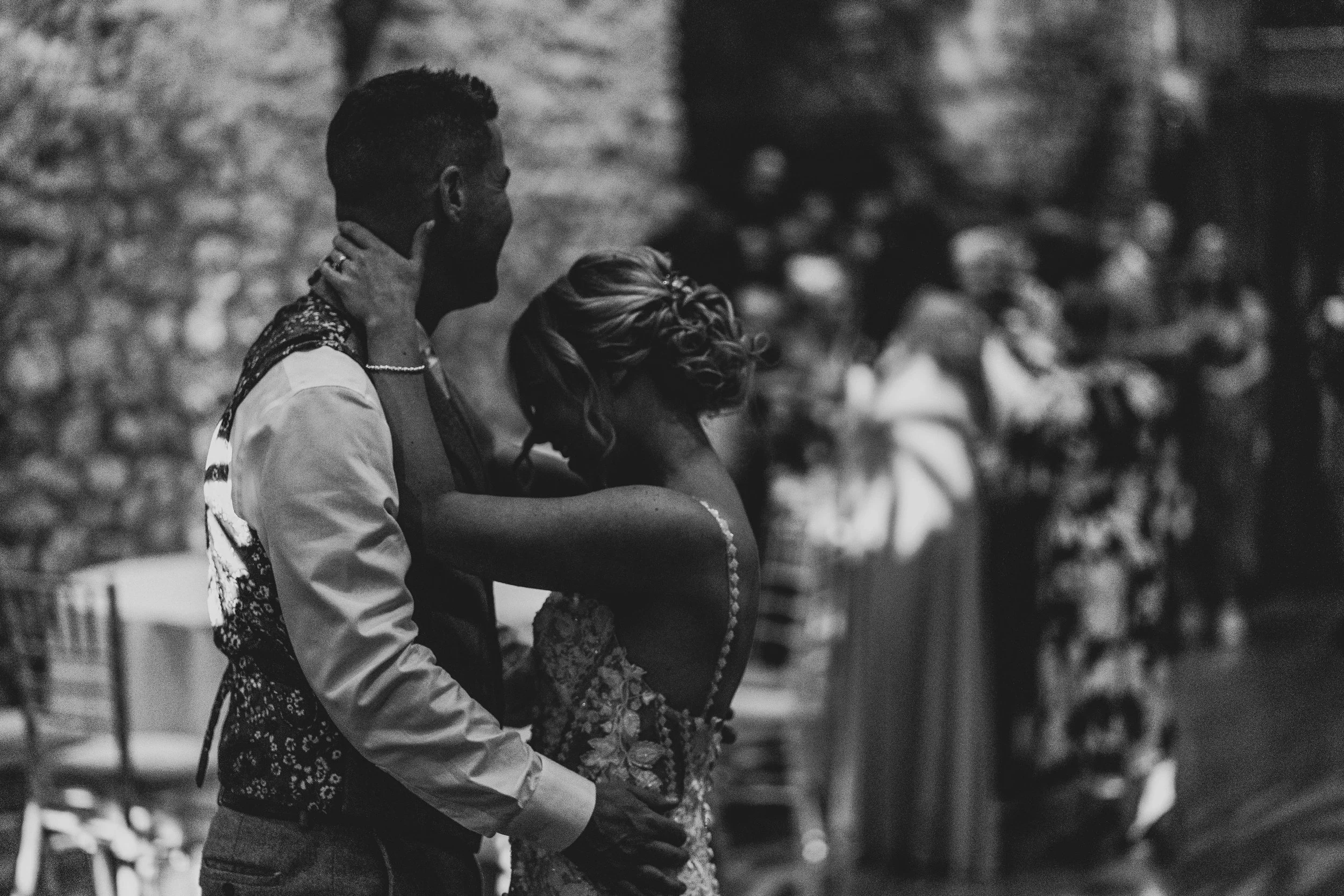 A black and white photo of a couple at their wedding, the groom in a suit with a patterned vest, and the bride in a strapless lace wedding dress, with their faces turned away from the camera, surrounded by wedding guests in the background.