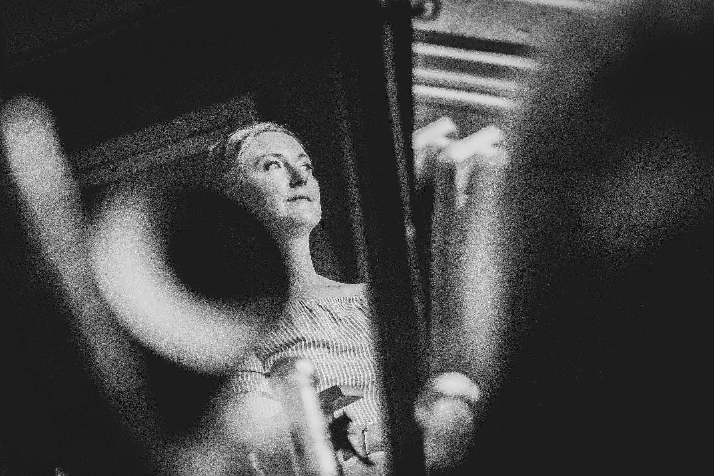 A black and white photo of a woman looking upward, viewed through a partially open door or window with blurred objects and furniture around her at Dillington House