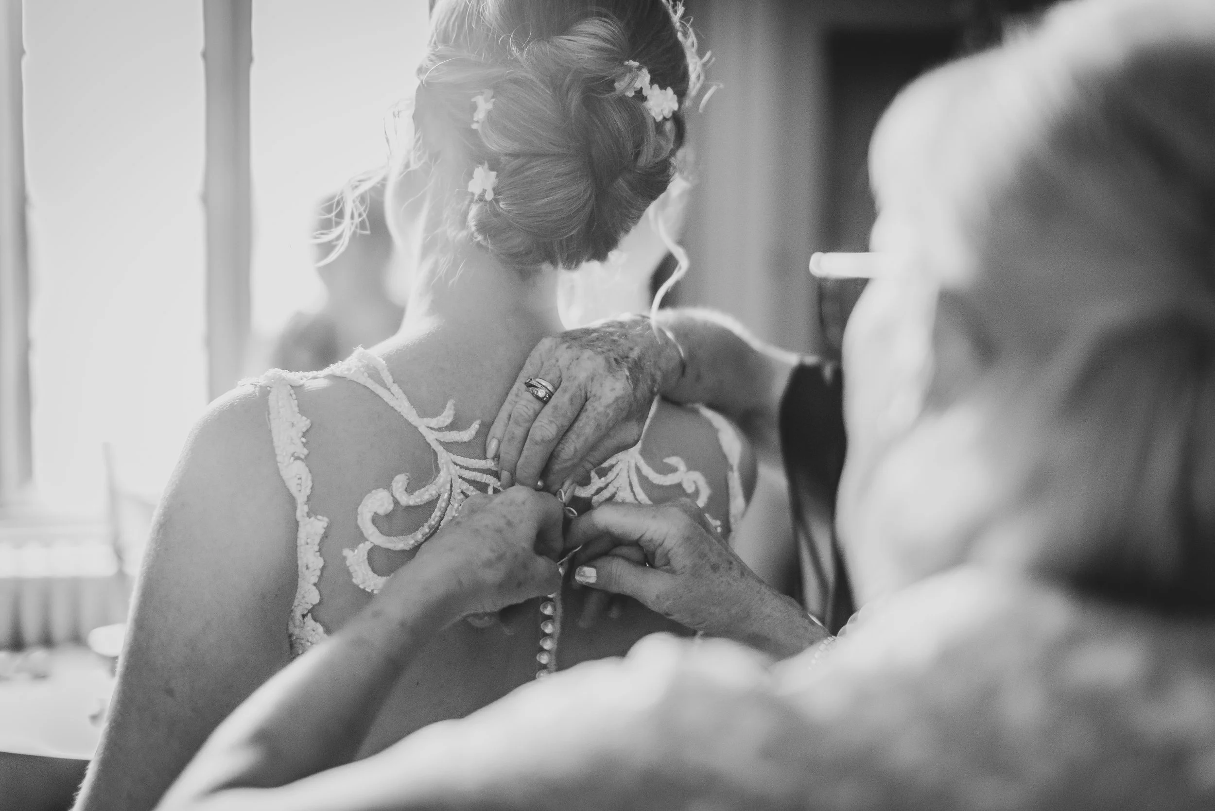 A woman in a wedding dress with lace details is being helped with her dress by her mother, in a black and white photo at Dillington House