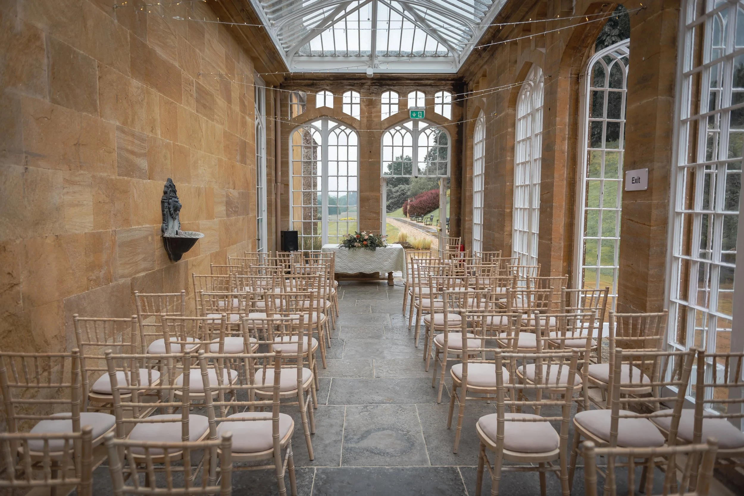An indoor wedding ceremony space with rows of beige chiavari chairs facing an altar table, large arched windows, a glass ceiling, and a scenic outdoor view at Dillington House