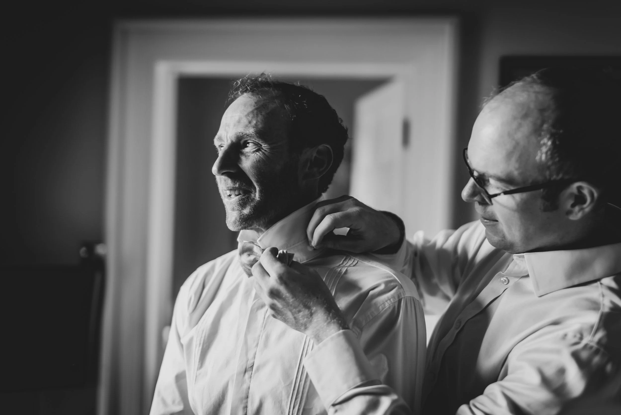 Two men in white shirts, one groom and one best man, adjusting the other's bow tie in a softly lit room, black and white photograph.