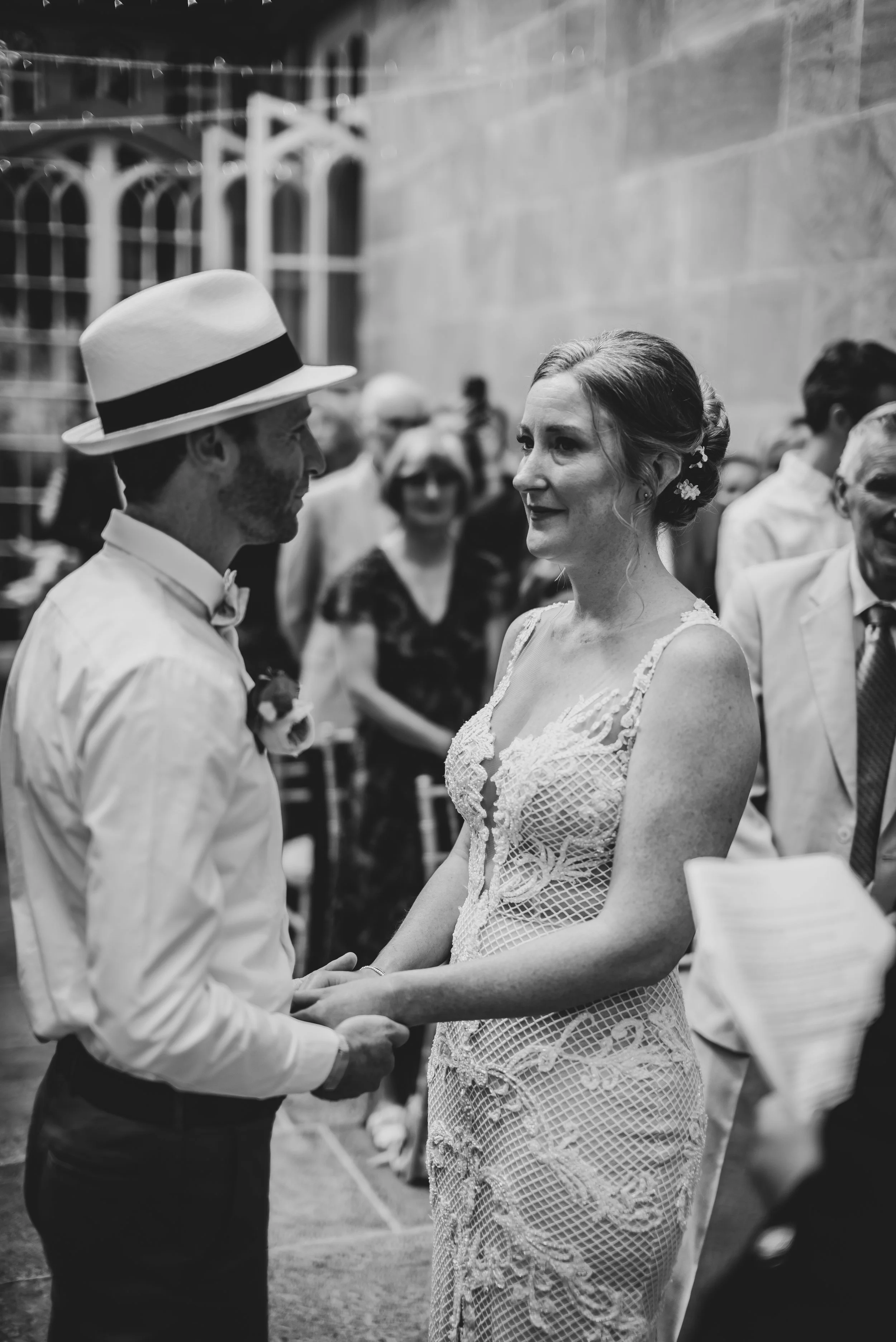 A black and white photo of a wedding ceremony with a man and woman holding hands and looking at each other, surrounded by guests at Dillington House