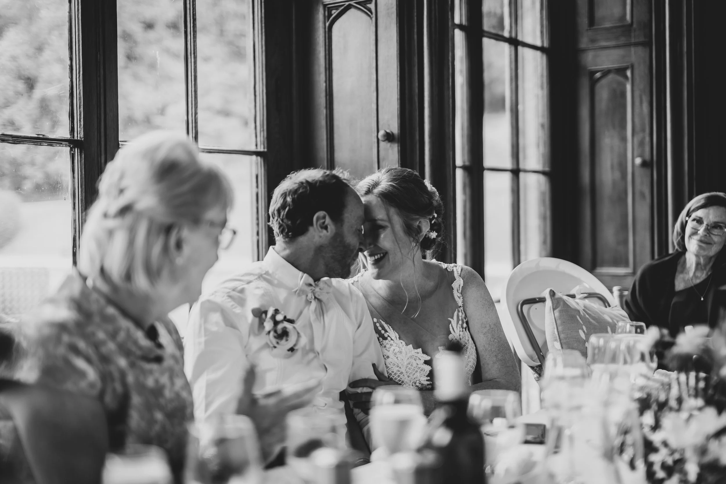South West wedding photographer captures a Black and white photo of a wedding reception with a bride and groom sitting close together, smiling with their foreheads touching, surrounded by seated guests.