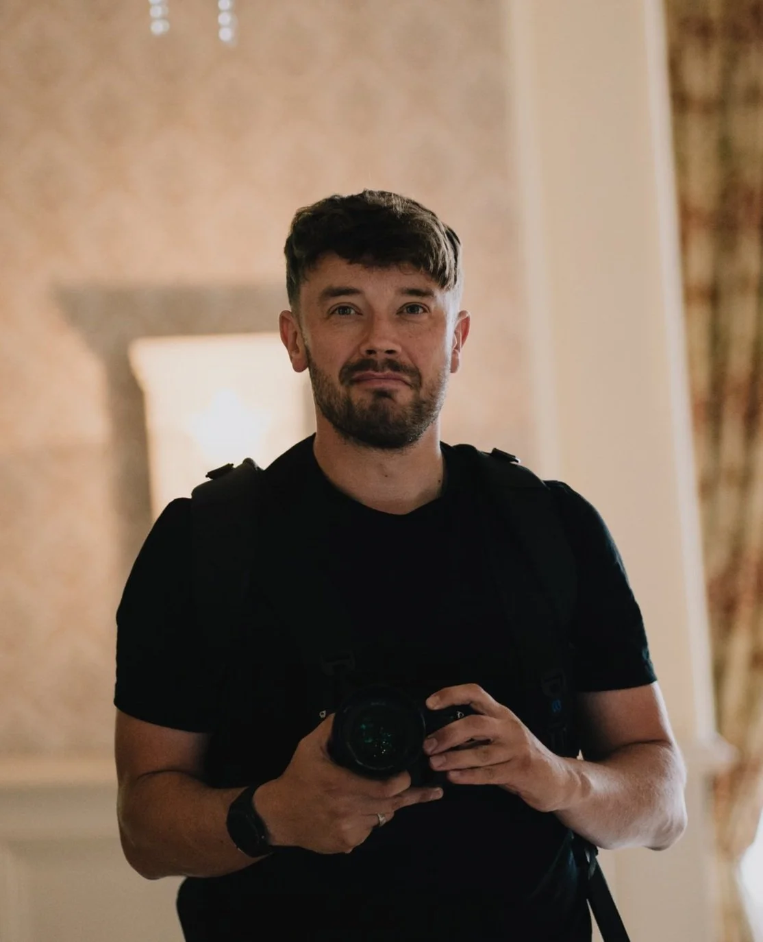 Mike Greenhill - Bristol Wedding Photographer with a camera in a dimly lit room. He is wearing a black shirt and a watch, with a backpack on his shoulders.