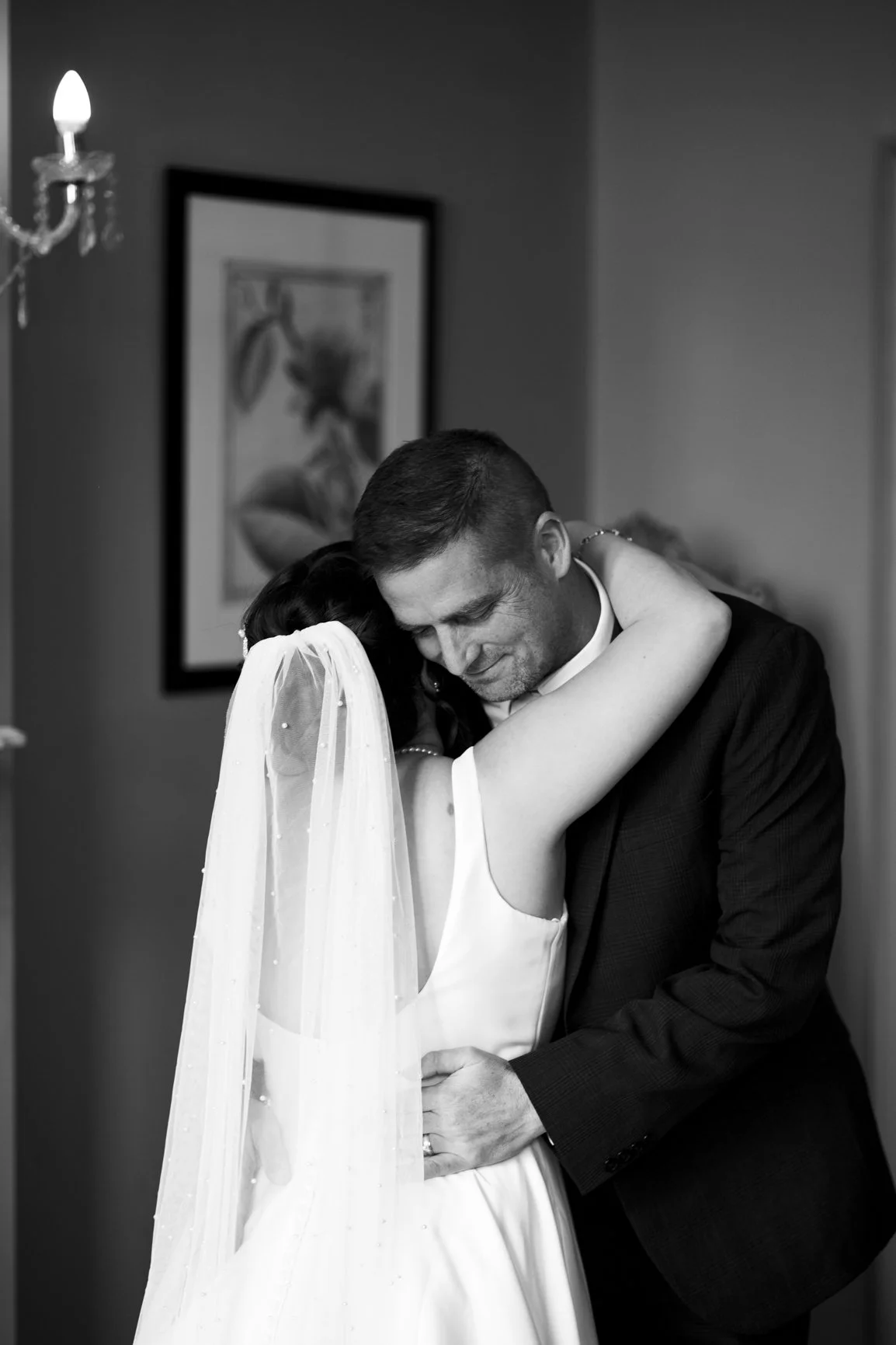 A black-and-white photo of a bride and groom embracing during their wedding, with the bride wearing a veil and sleeveless dress and the groom in a dark suit.