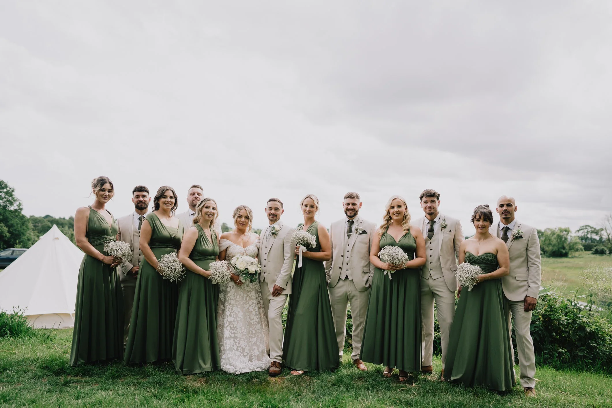 A wedding party of 12 people, including the bride in a white lace dress and 11 bridesmaids and groomsmen, standing outdoors on grass with trees and a partly cloudy sky in the background. The women are wearing long green dresses, and the men are in light gray suits with matching ties.