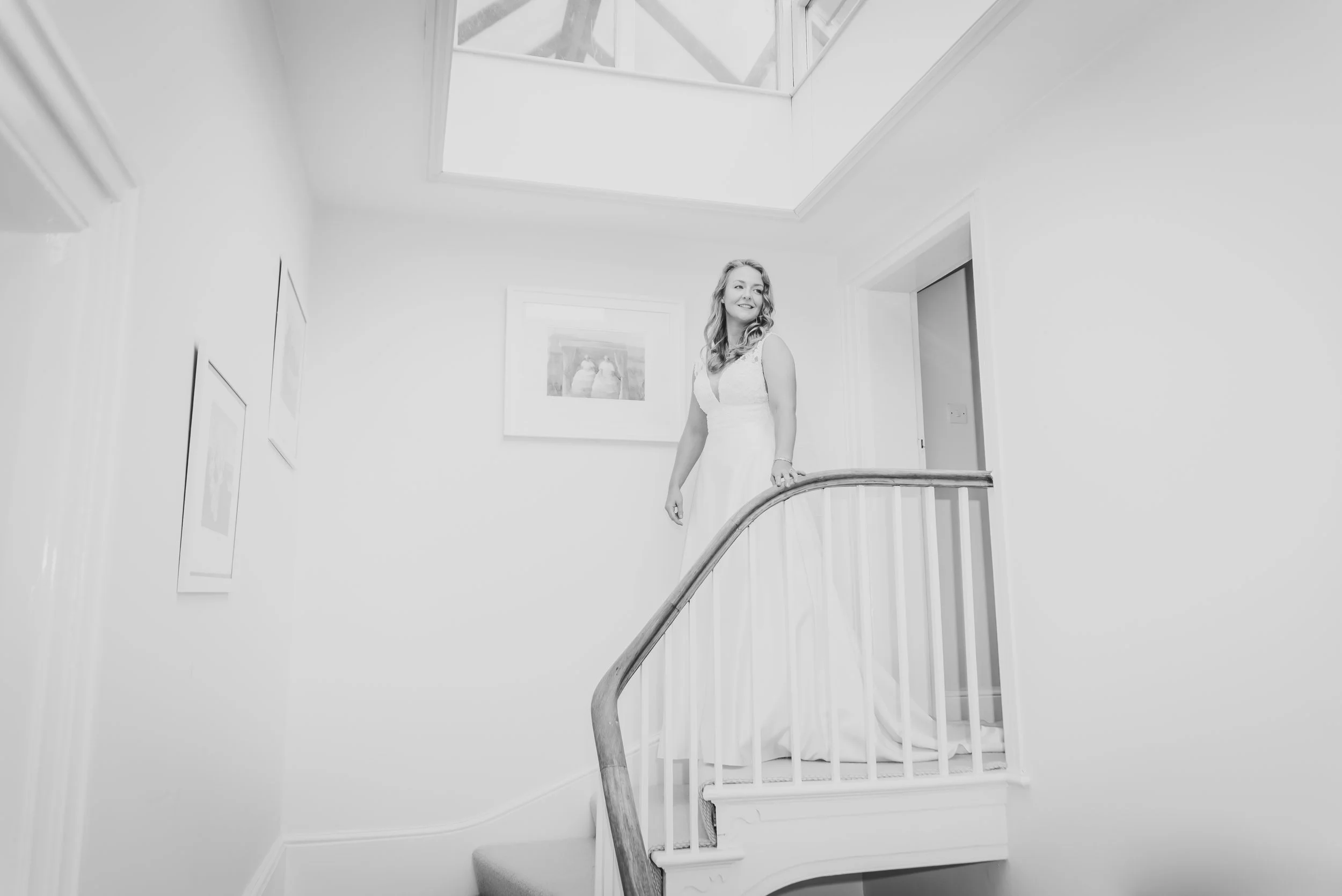A bride in a wedding dress standing on a staircase in a bright, white-walled interior with framed artwork and a skylight ceiling during prep captured by Clifton Wedding Photographer