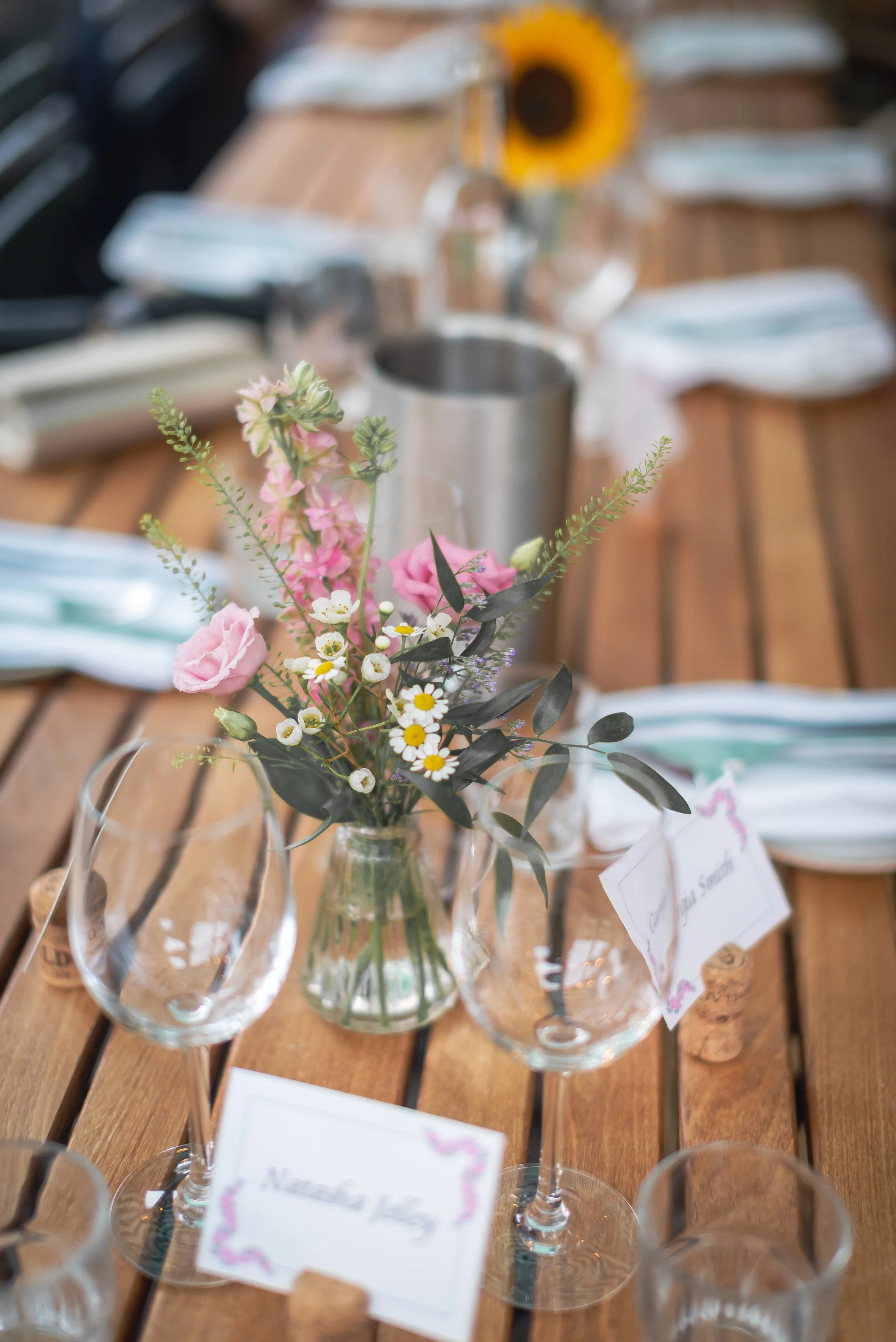 A wooden table decorated with pink and white flowers in a glass vase, wine glasses, a metal cup, and place cards at an outdoor event at Snobbys in Bristol