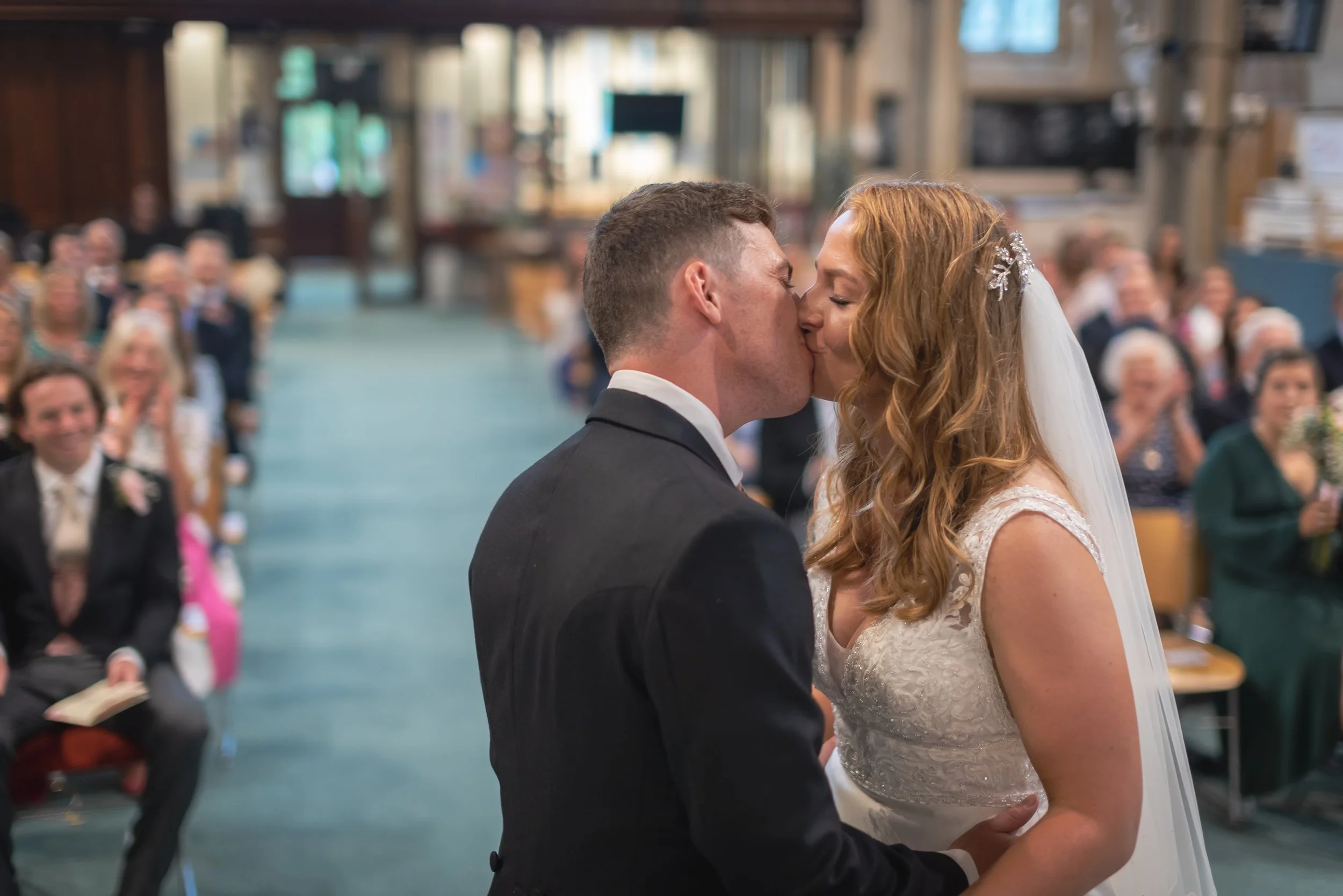 A bride and groom share a kiss during their wedding ceremony inside a church, with guests seated and watching in the background captured by Clifton Wedding Photographer