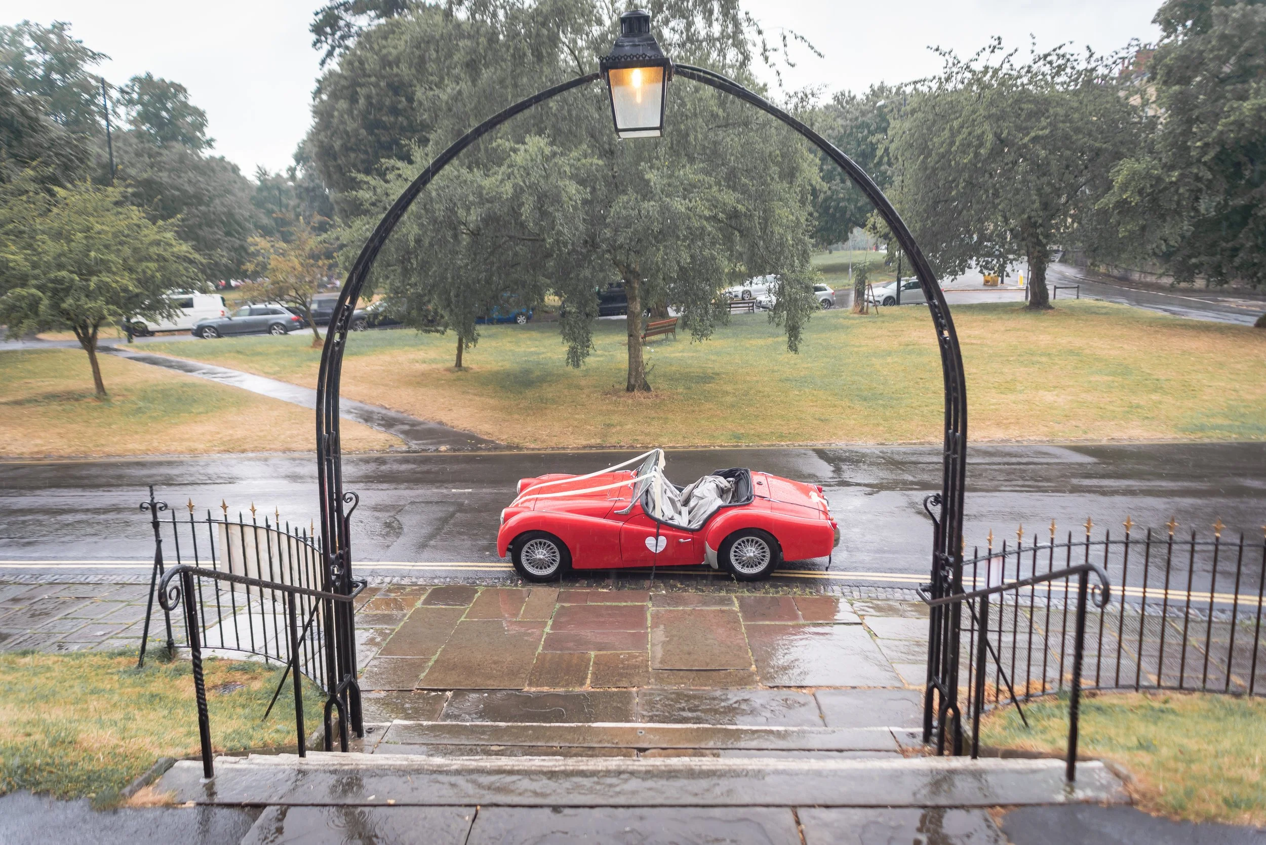 Rainy day view through an arched iron gate showing a small red vintage convertible car parked on a wet street. The background features a grassy park, trees, and a row of parked cars along the street after the wedding ceremony in Clifton, Bristol.
