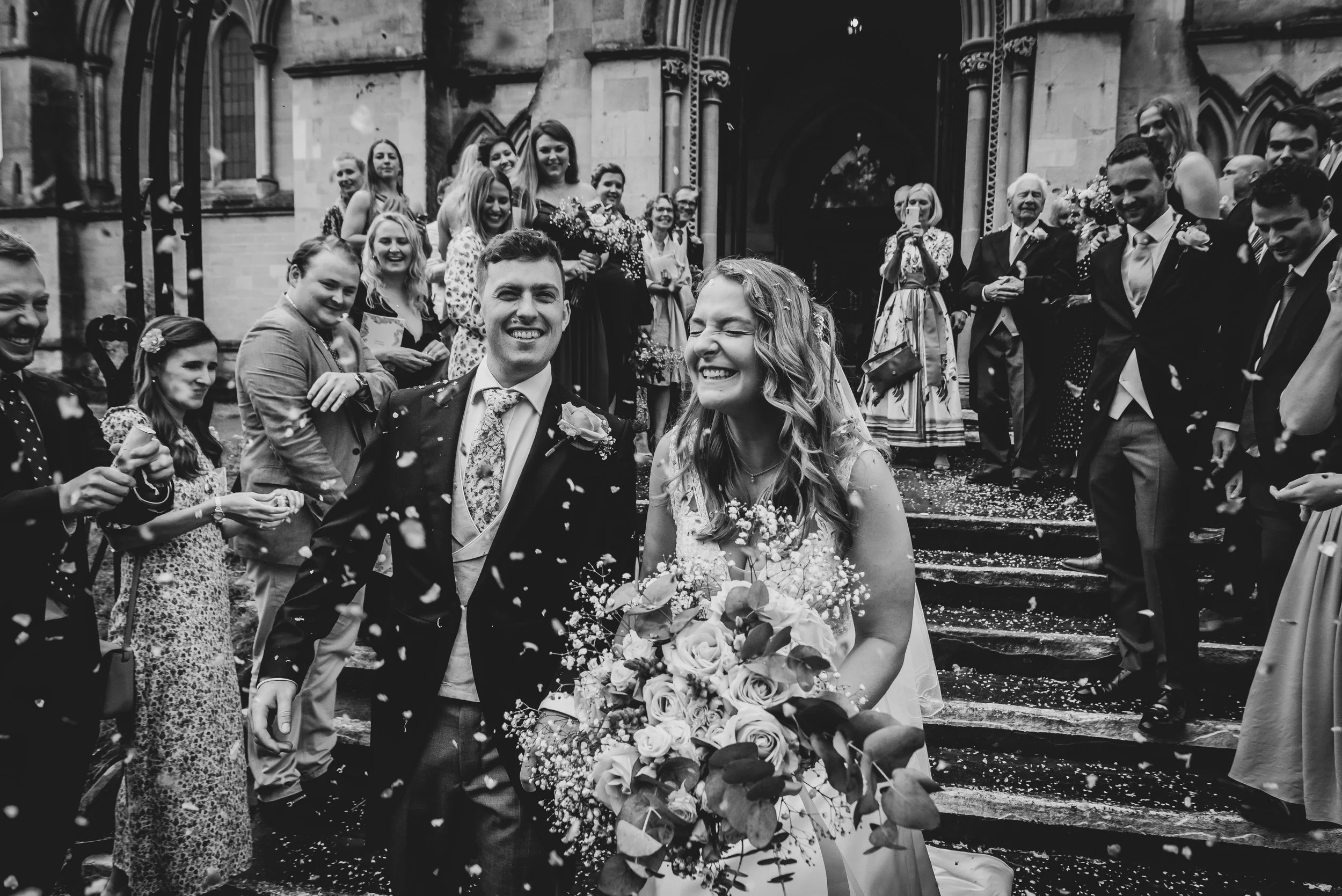 Black and white photo of a newlywed couple exiting a church surrounded by friends and family with confetti falling in Clifton, Bristol.