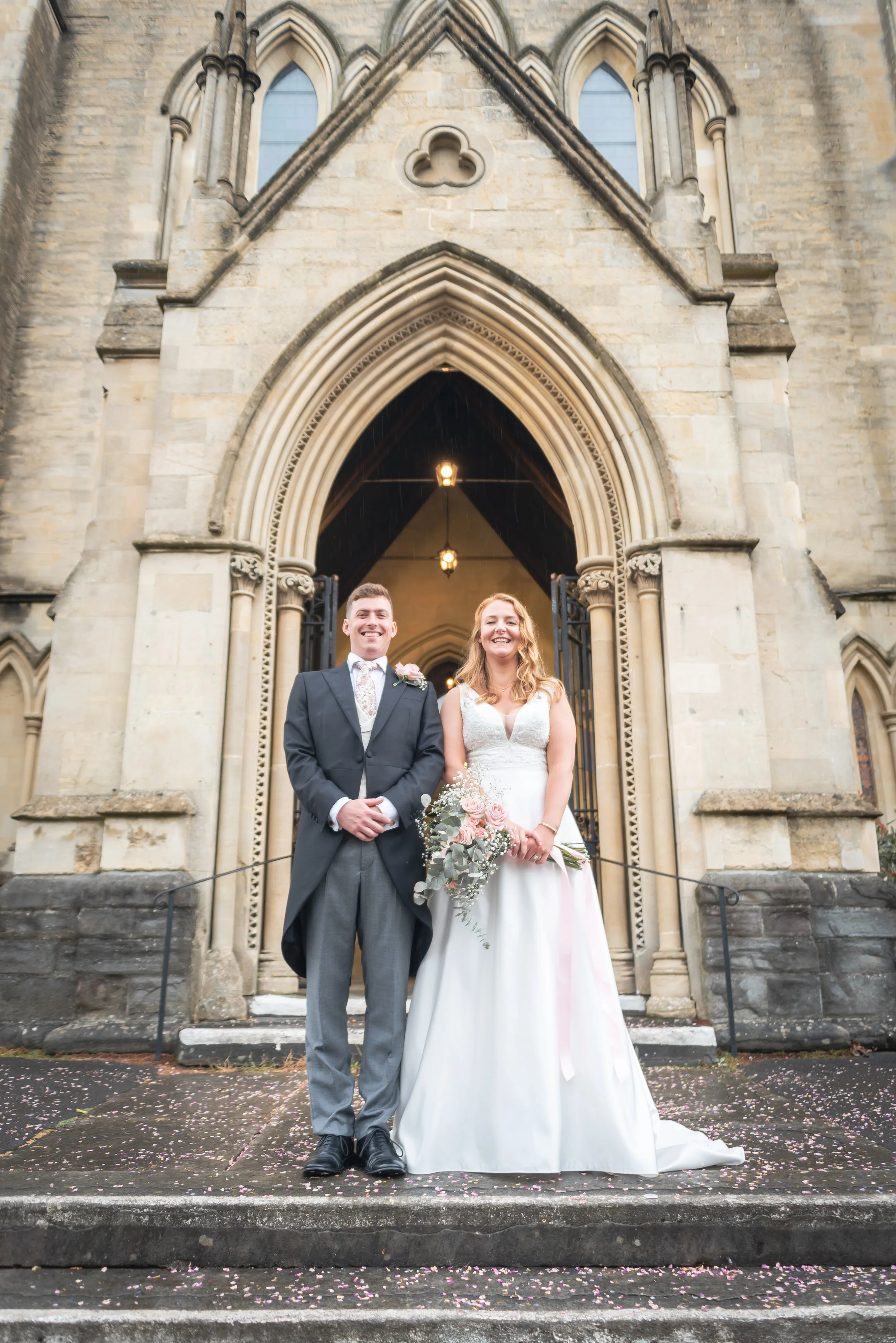 A newlywed couple stands on the steps outside a historic stone church, smiling and holding flowers, on a wedding day captured by Bristol Wedding Photographer