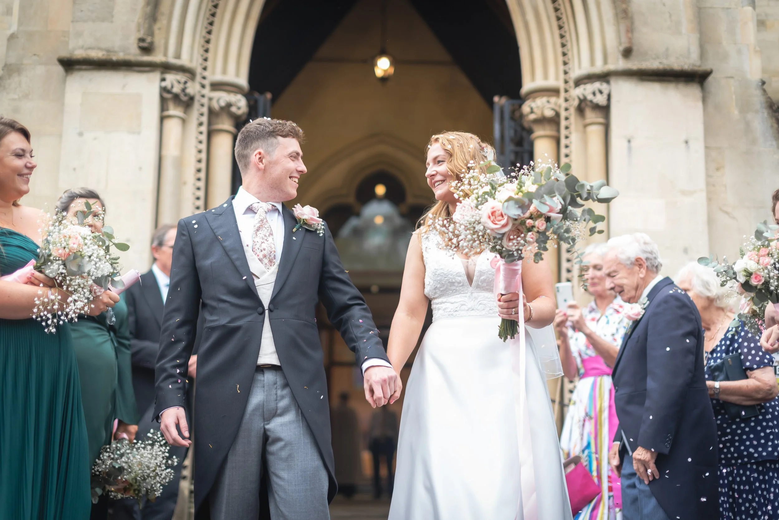 Bride and groom smiling and holding hands outside a church, surrounded by wedding guests, with confetti falling around them for a wedding photographer in Bristol.