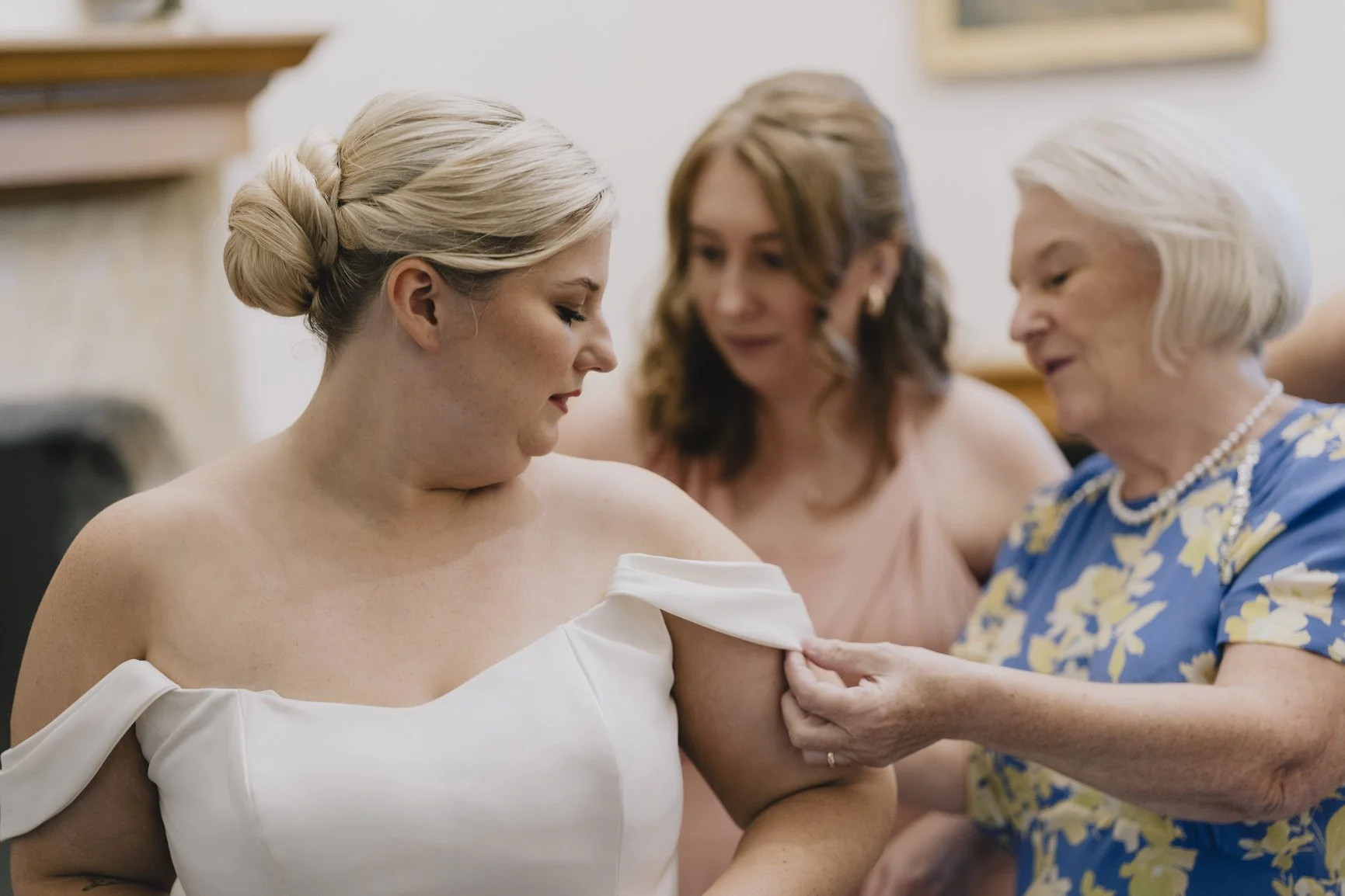 A bride wearing an off-the-shoulder white wedding dress is being helped with her dress by an elderly woman in a blue floral dress, while another woman in a pink dress looks on.