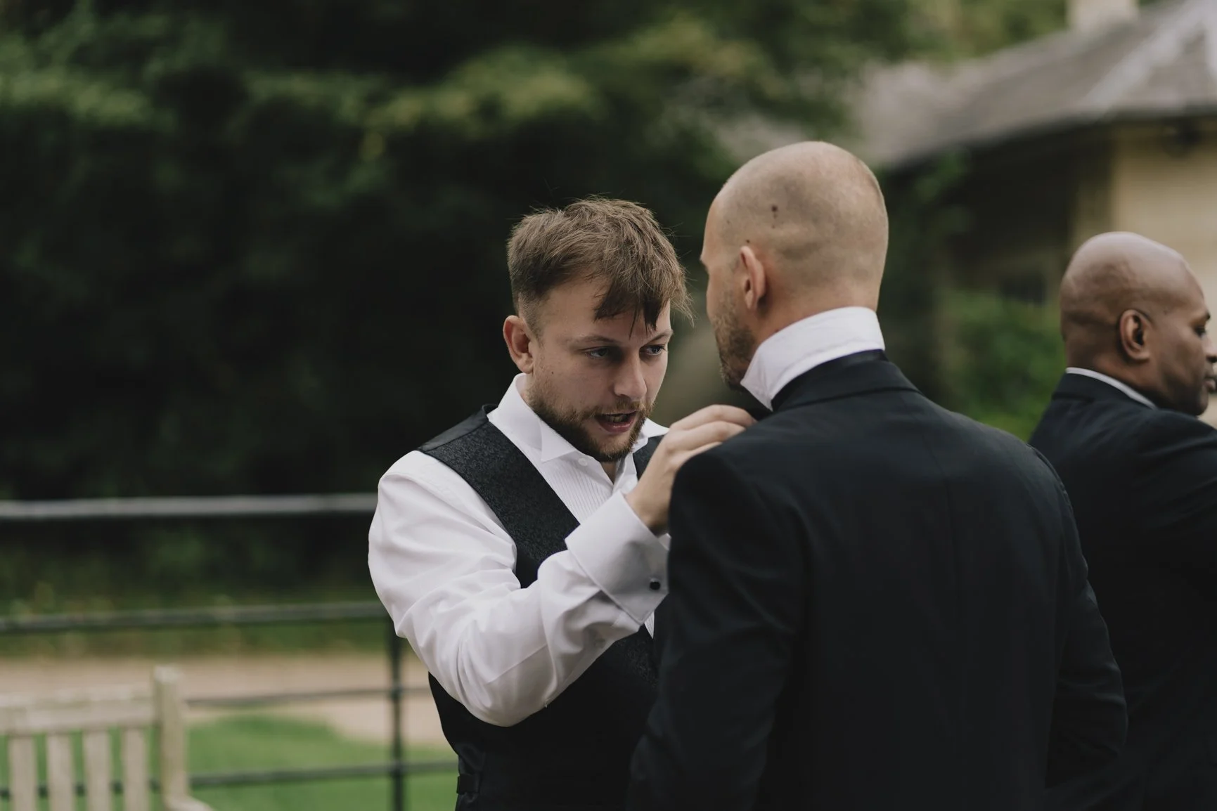 Three men dressed in formal suits, two of them black and white, groomed, talking outdoors with trees and a rustic building in the blurred background at Orchardleigh Estate