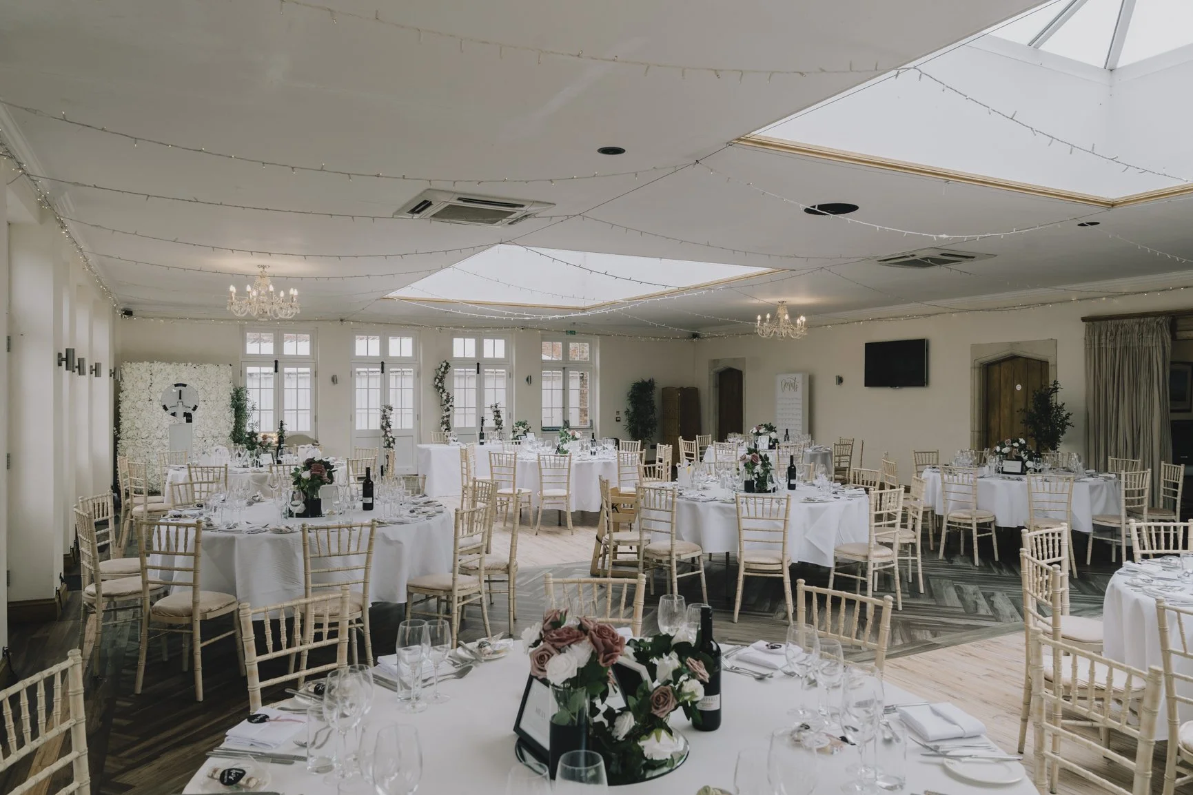 Elegant banquet hall at Elmay Park decorated for an event with white round tables, gold chairs, floral centerpieces, and string lights on the ceiling.