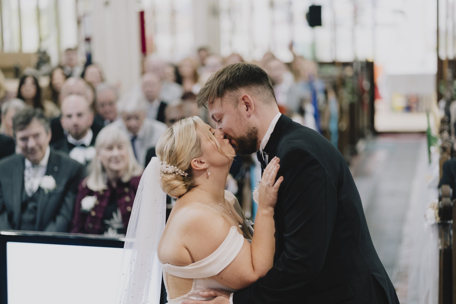 A bride and groom share a kiss during their wedding ceremony, surrounded by seated guests in an indoor setting.