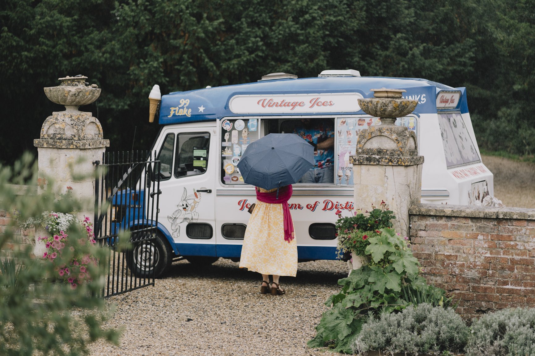 A woman holding a black umbrella stands in front of a vintage ice cream truck, decorated with cartoon characters and colorful signs, on a gravel path, surrounded by a garden with flowers and bushes.