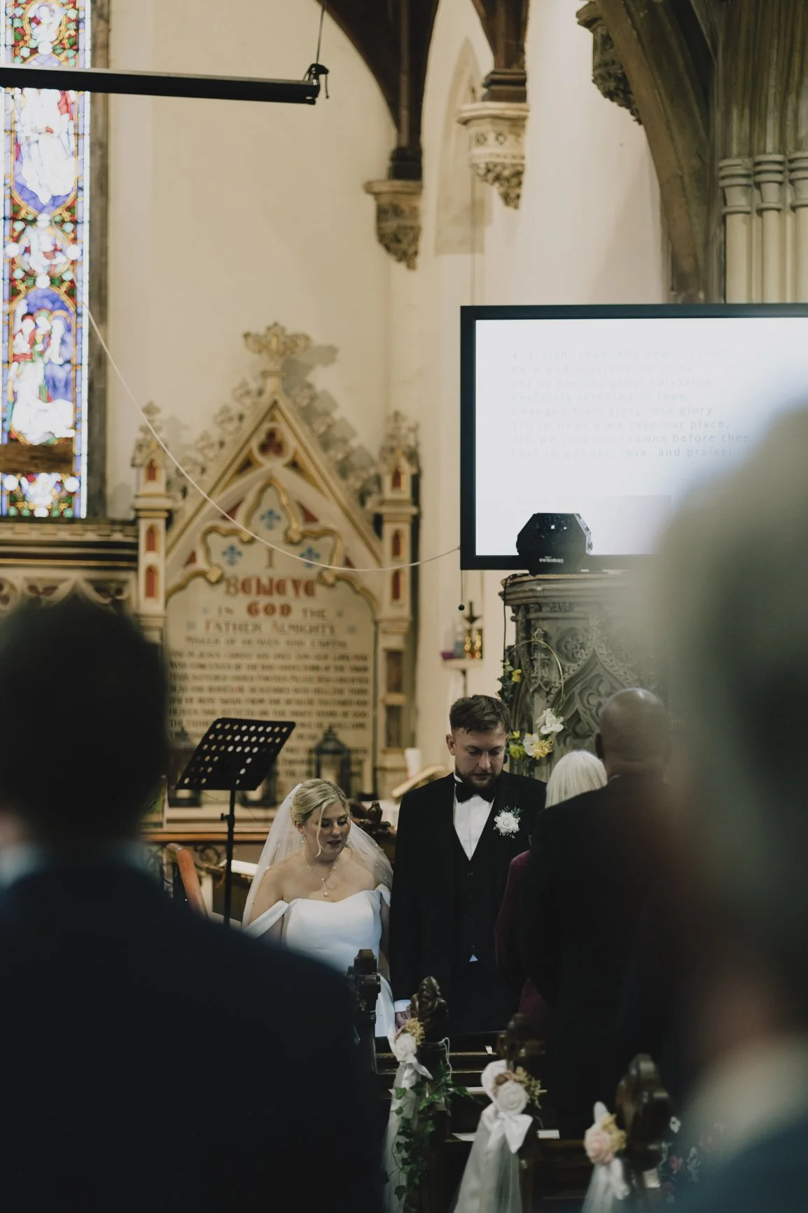 A wedding ceremony taking place inside a church with stained glass windows. The bride and groom are standing at the altar, with the bride in a white wedding dress and the groom in a black tuxedo. Guests are visible in the foreground and a large screen with text is displayed to the right.