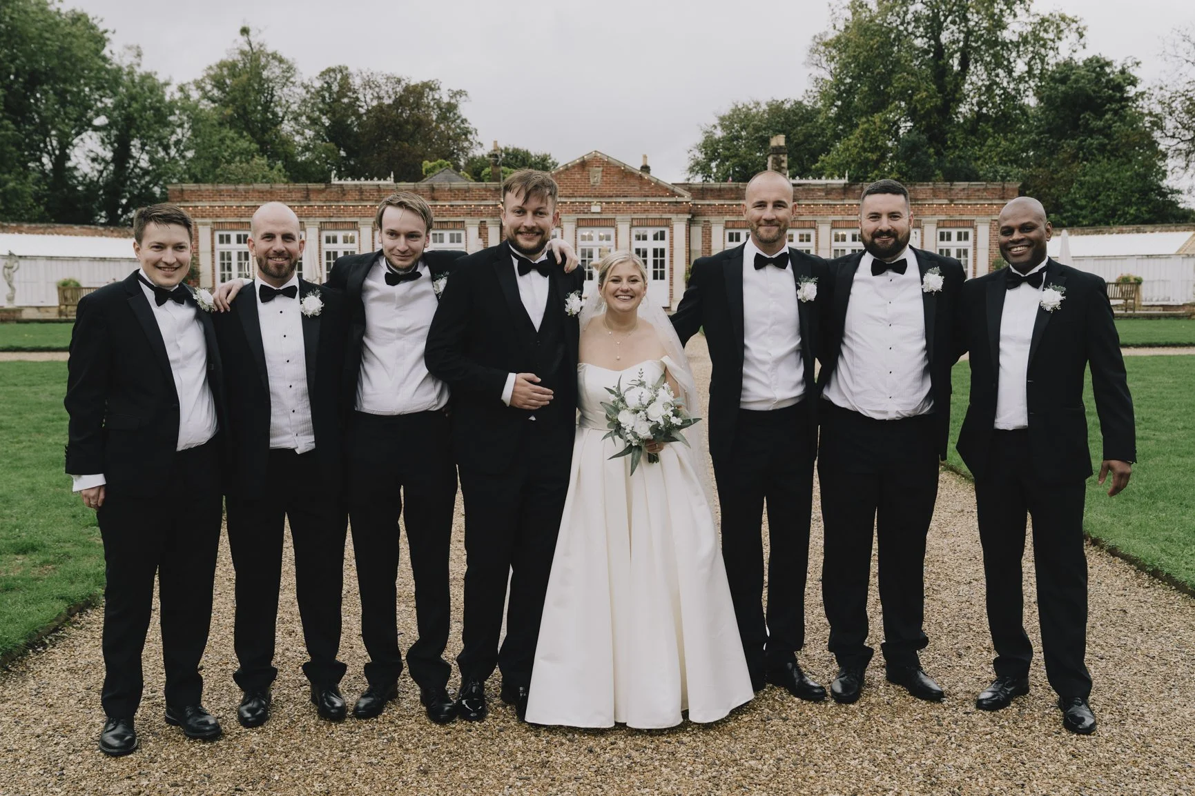 Wedding group photo outdoors with a bride in a white gown holding a bouquet, surrounded by seven groomsmen in black tuxedos. They are standing on a gravel path with a Elmhay Park and trees in the background.
