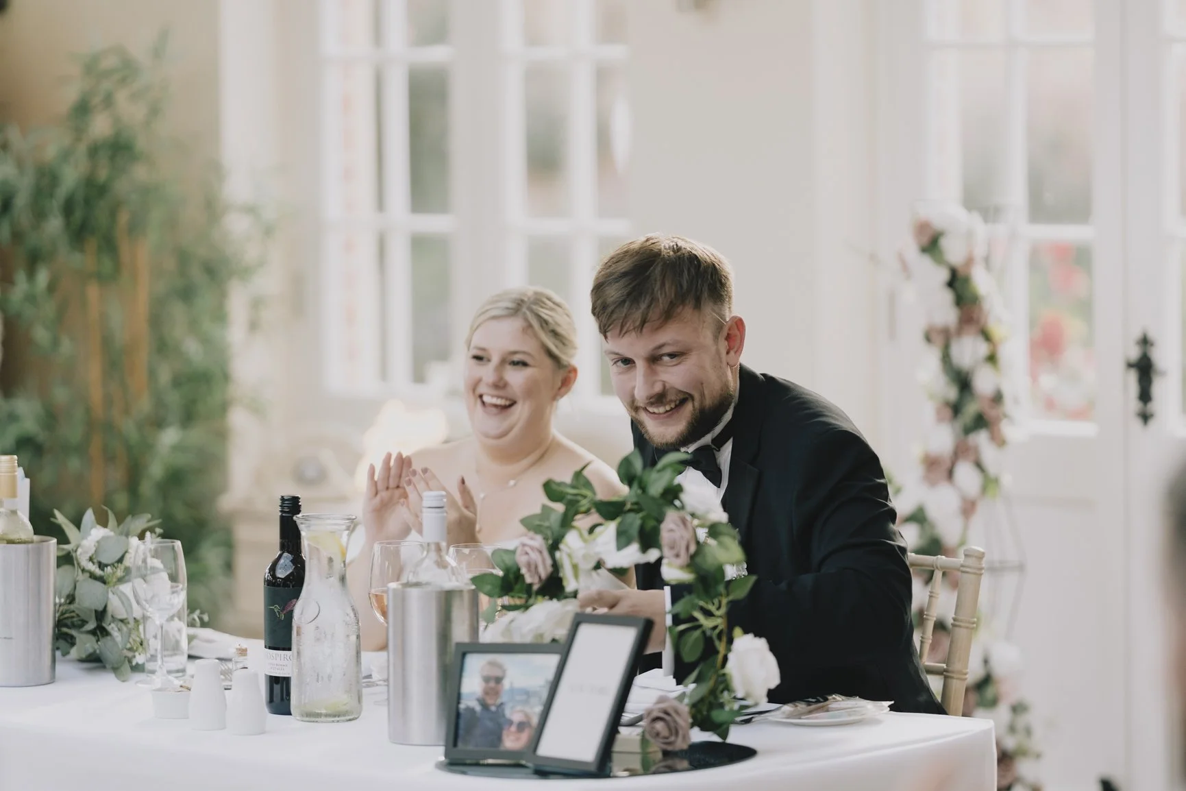A smiling bride and groom at their wedding reception seated at a decorated table with wine bottles, glasses, and floral arrangements in a bright room with large windows taken by Somerset Weddin Photographer.
