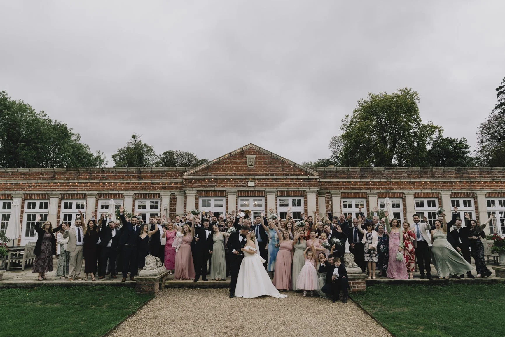Group of wedding guests gathered outside with white-framed windows, celebrating a wedding with many people raising their hands in the air. The bride and groom are in the center, with the bride in a white dress and the groom in a dark suit, sharing a kiss.