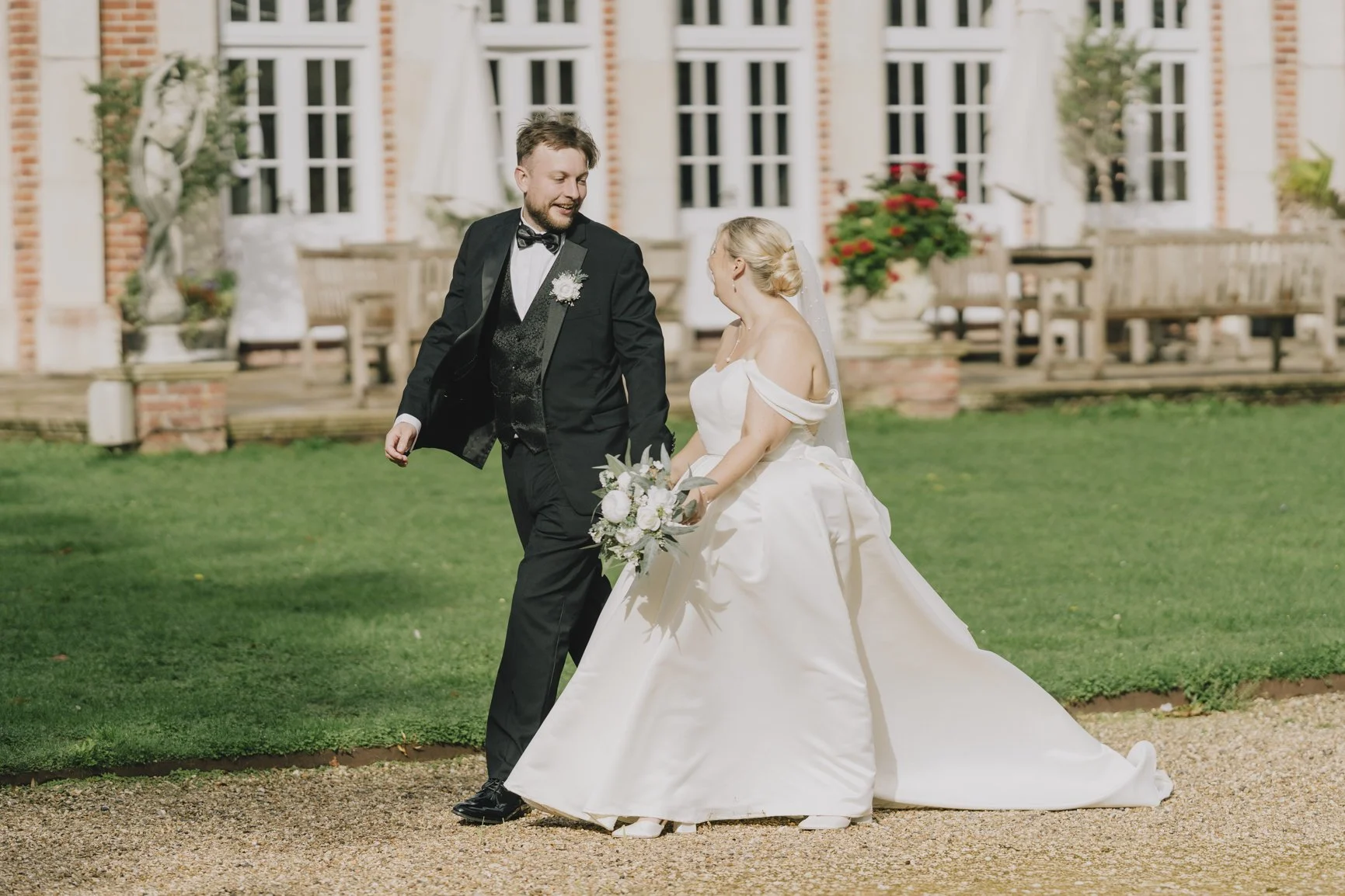 A bride and groom in wedding attire holding hands and smiling on a garden lawn in front of a house with white-framed windows and urn planters. The bride is wearing a white off-the-shoulder wedding gown and holding a bouquet.