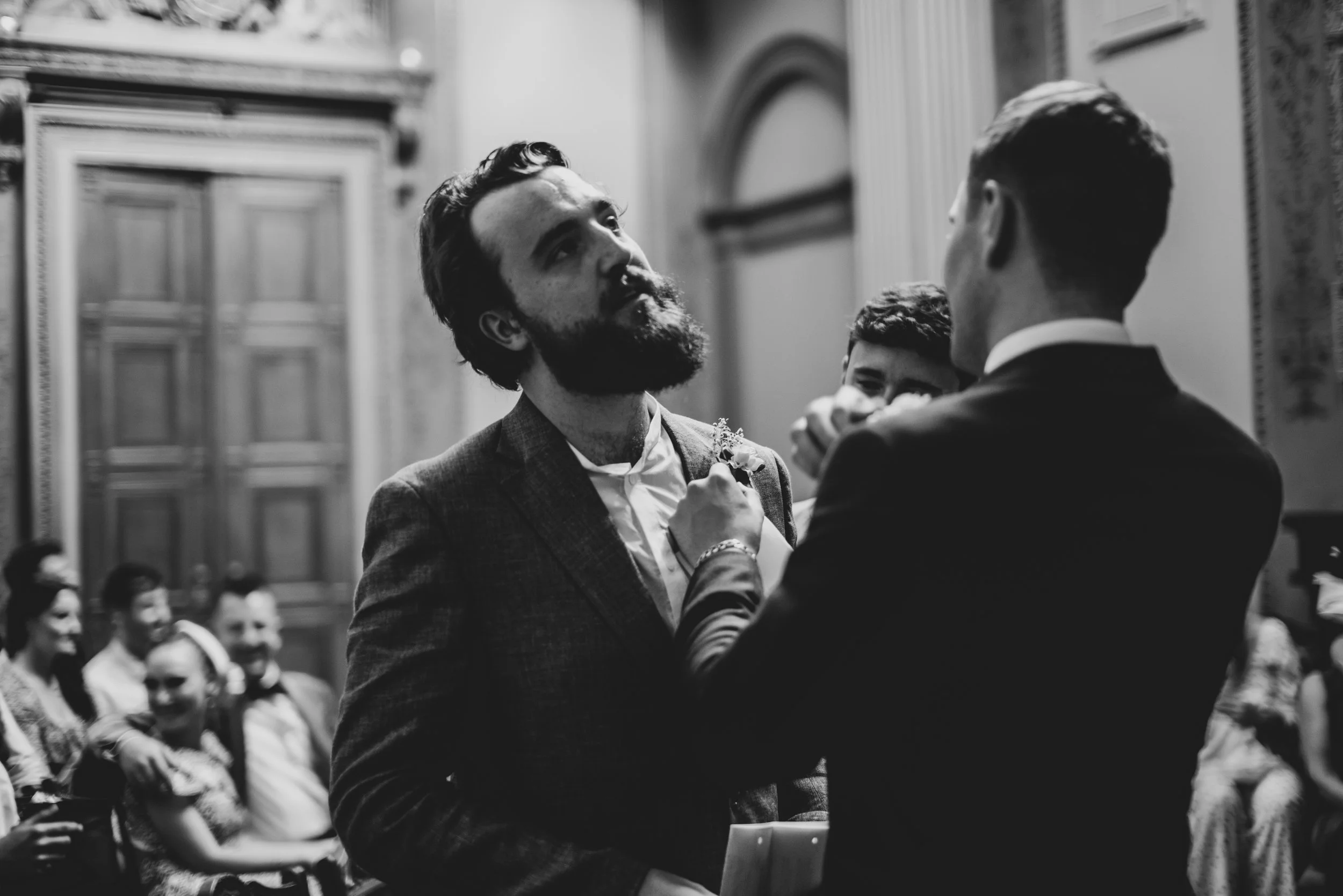 Black and white photo of a wedding ceremony at Bristol Registry Office with two grooms facing each other, one fixing the other's boutonniere, with guests seated in the background.