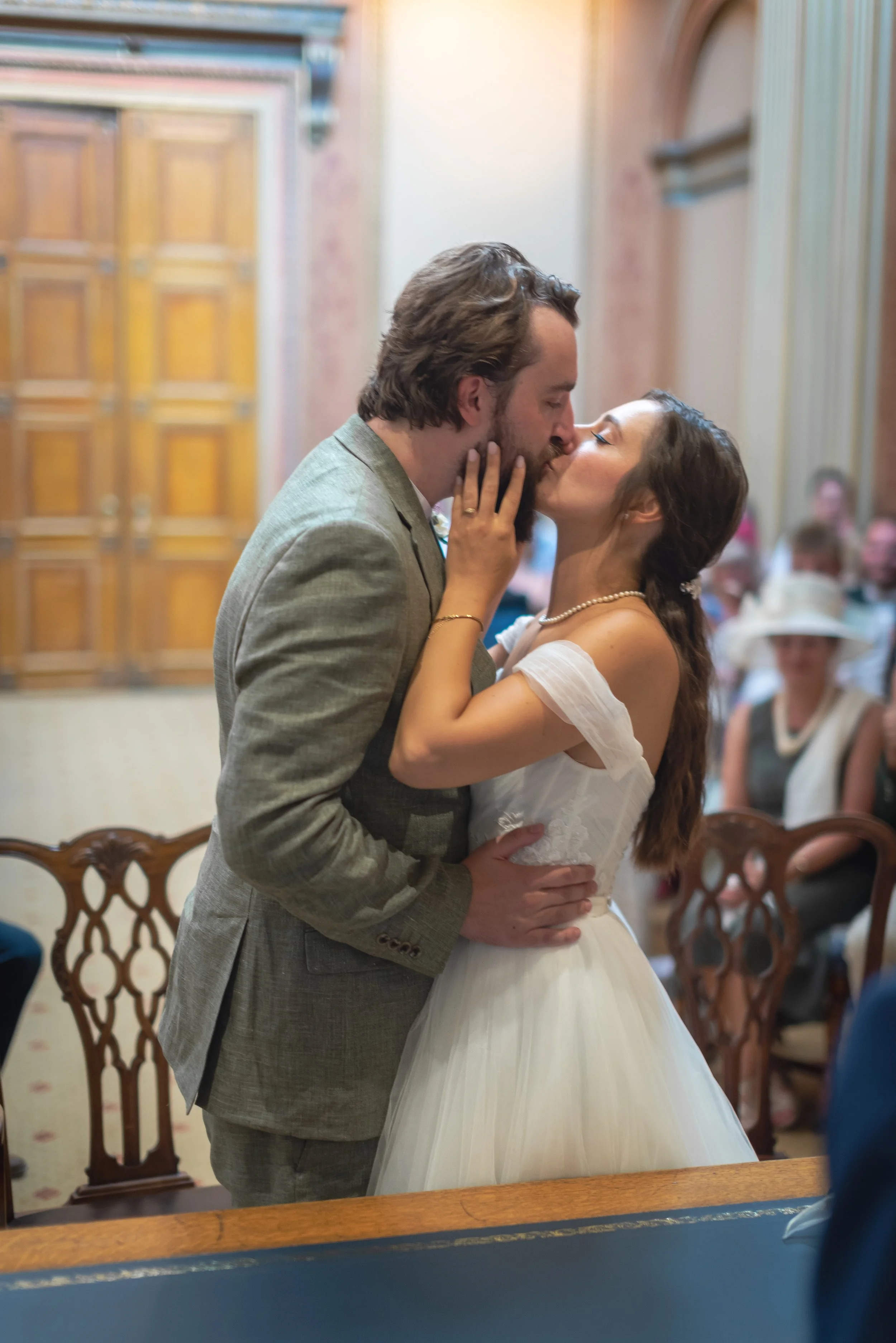 A newlywed couple sharing a kiss during their wedding ceremony in a grand hall, with guests seated in the background captured by Bristol Wedding Photographer