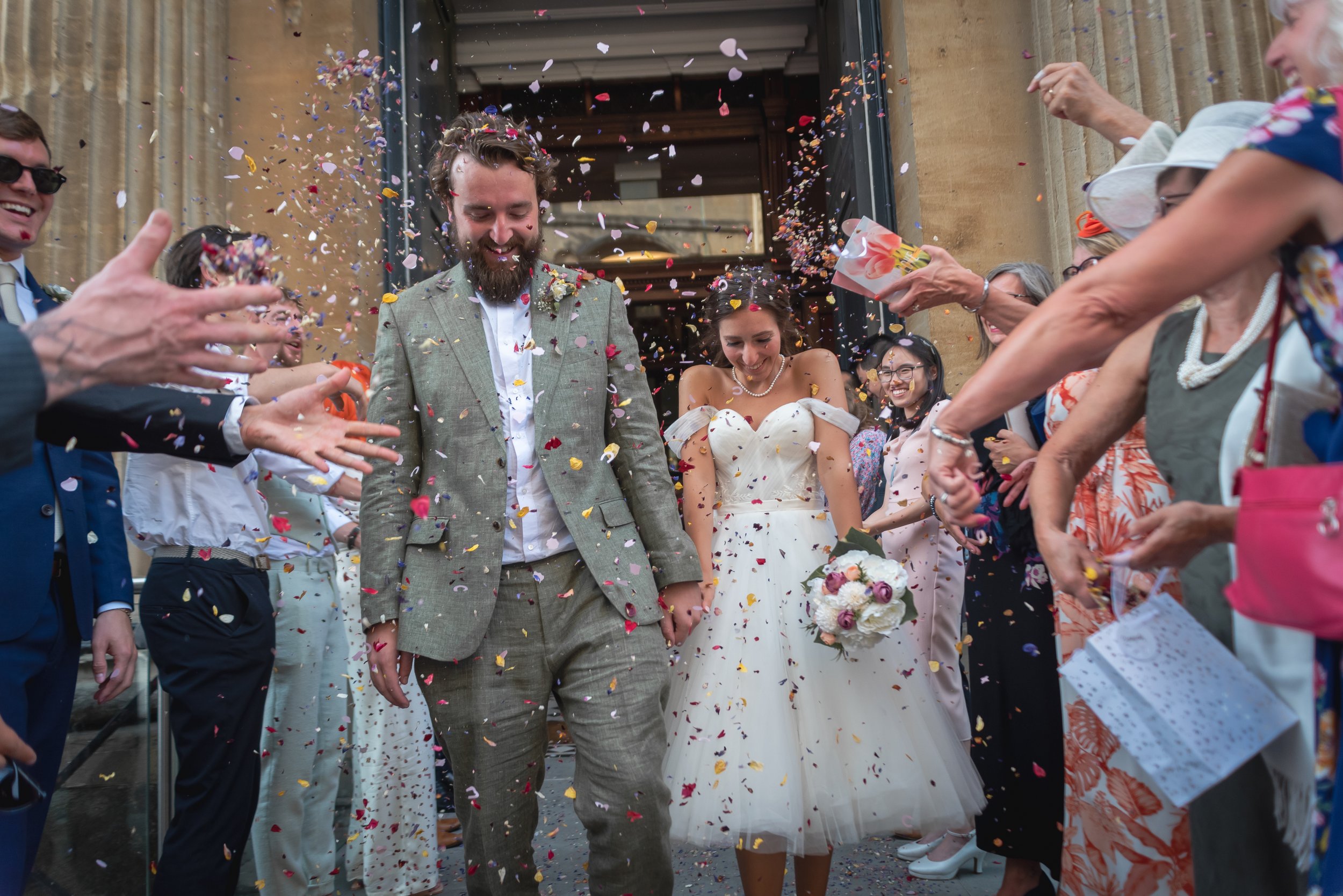 Bristol Wedding Photographer captures newlywed couple being showered with colorful confetti by friends and family outside a building.