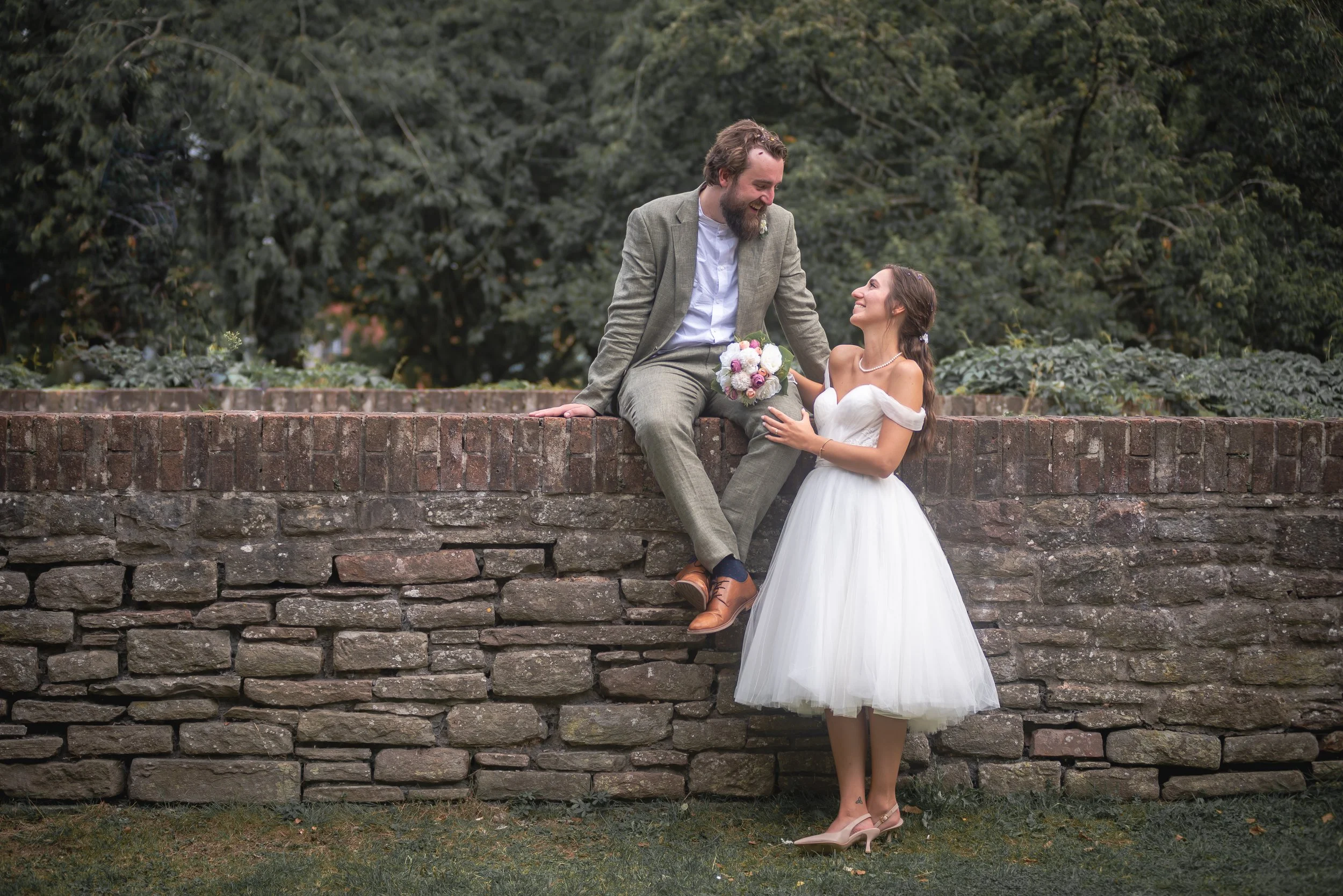 A bride and groom on a stone wall outdoors, smiling and looking at each other. The groom, sitting on the wall, is wearing a light gray suit with a white shirt in Castle Park, Bristol captured by wedding photographer