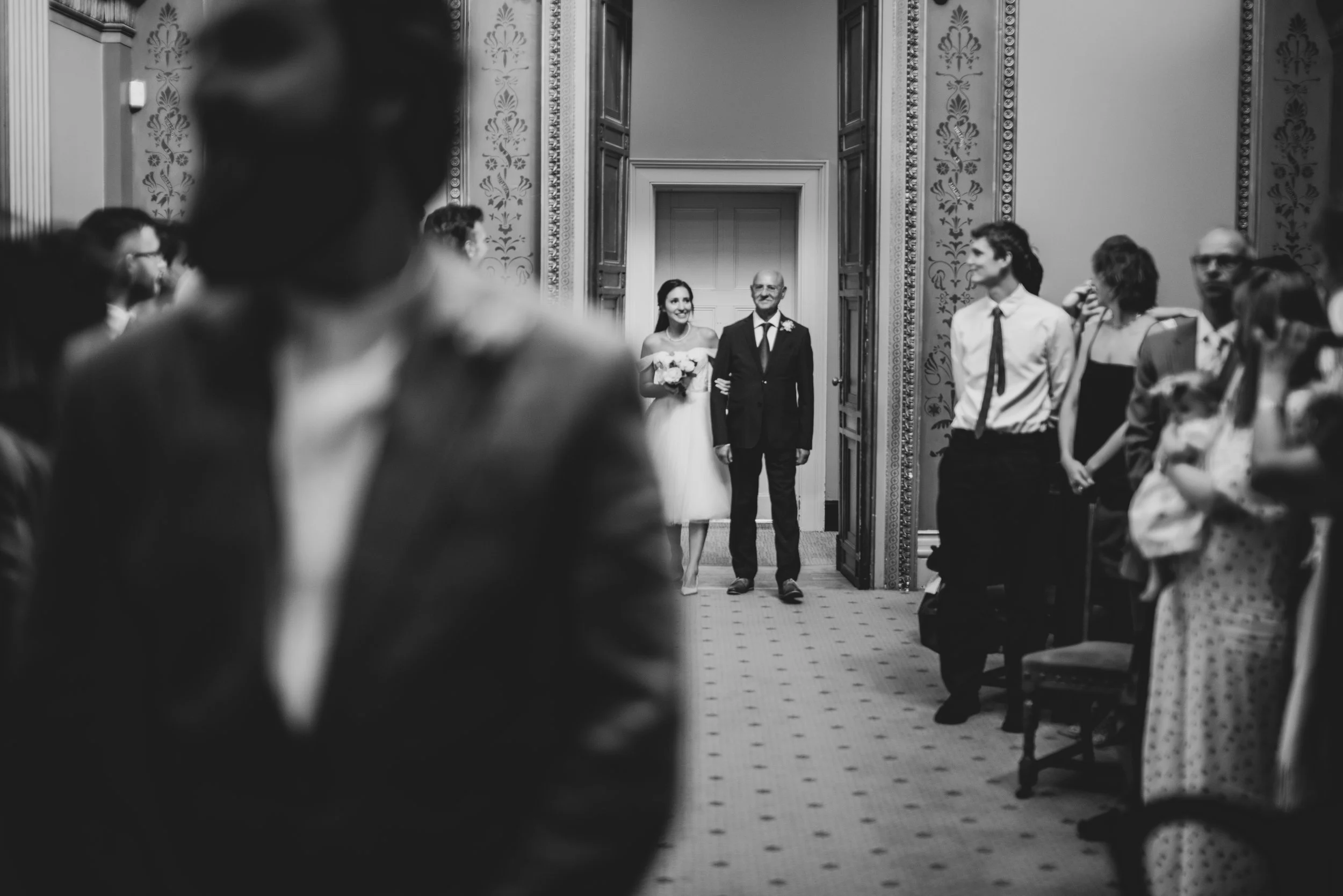 Black and white photo of a bride walking down the aisle with her father, at a wedding ceremony. The bride is holding a bouquet and smiling. Guests are seated on either side of the aisle, some standing and watching as wedding photographer waits.