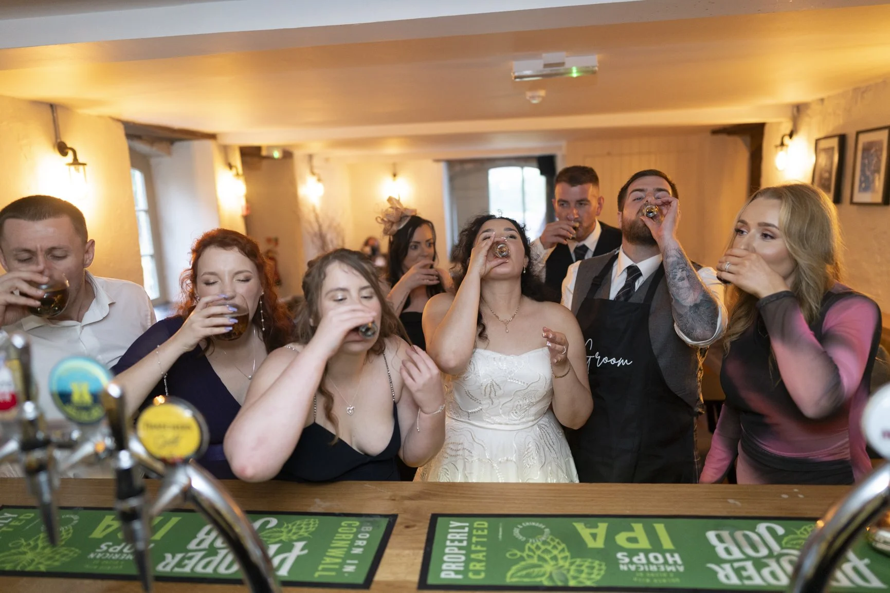 Guests at Priston Mill in Bath doing shots with Bride and Groom as Bath Wedding Photographer takes photo from behind the bar