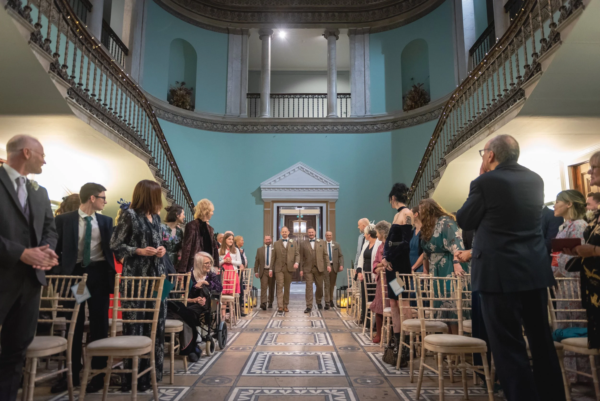 Andy and Issac walking joyfully down the aisle, flanked by their two best men, in the ceremony room at Leigh Court.