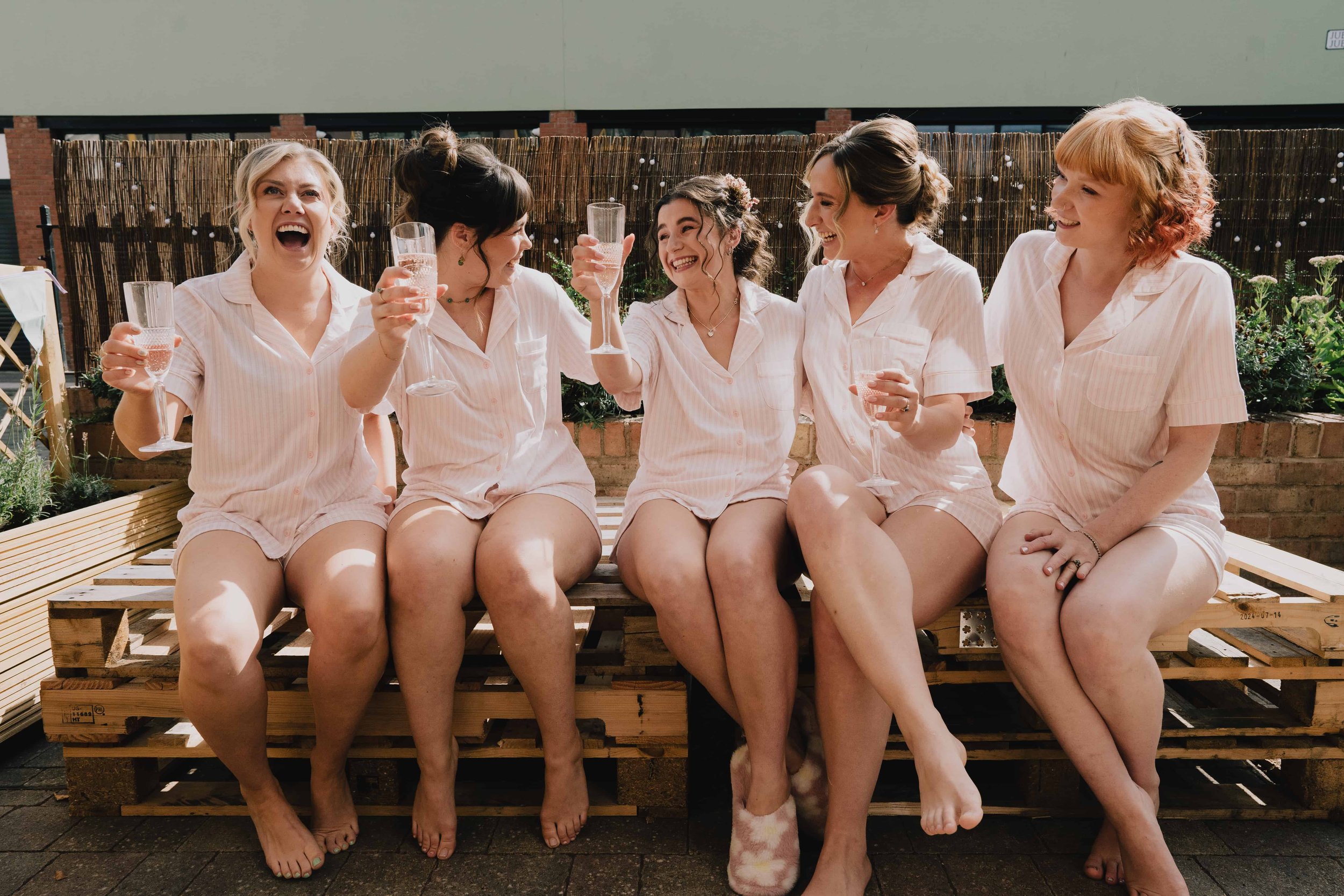 Six women wearing matching light pink pajamas sitting on a wooden pallet bench, celebrating with glasses of champagne in an outdoor setting ready for a wedding in Bristol taken by Bristol Wedding Photographer