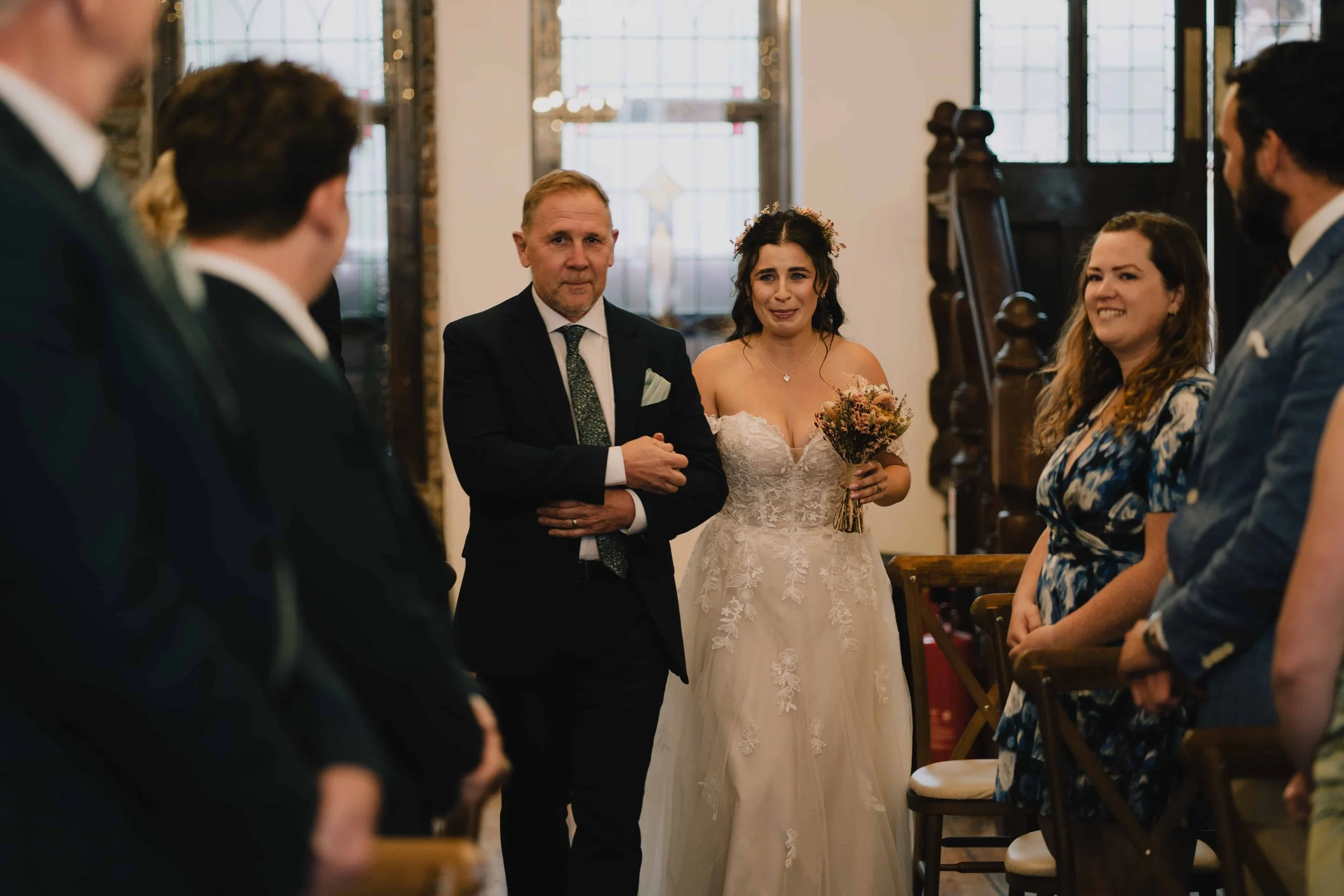 A bride in a wedding dress holding a bouquet crying while walking down the aisle, accompanied by a man in a suit. Guests stand on either side, watching the emotional moment in a decorated Radnor Rooms, Bristol.