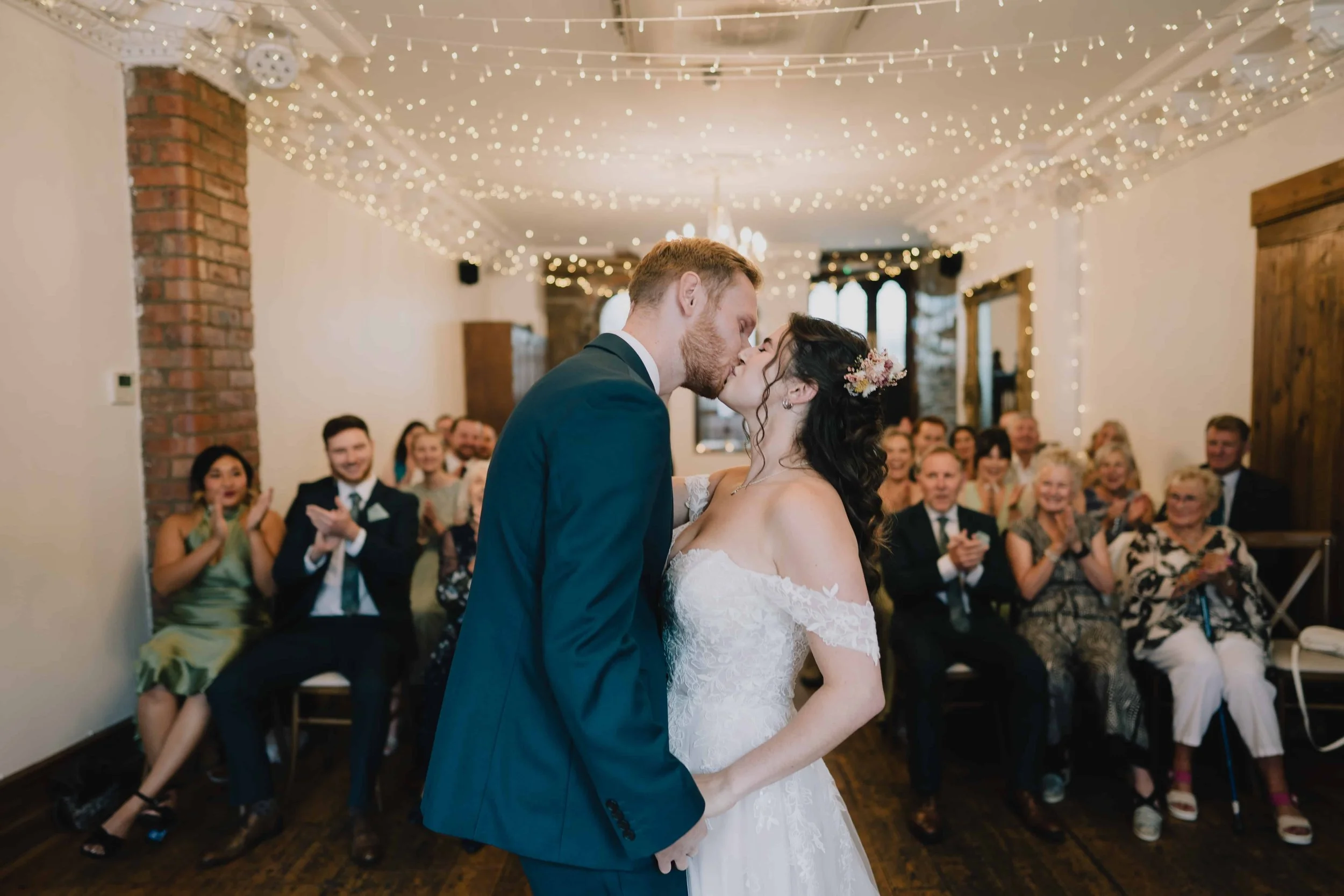 A newlywed couple kissing during their wedding ceremony in Radnor Rooms Bristol. The bride is in a white off-shoulder wedding dress with flowers in her hair.