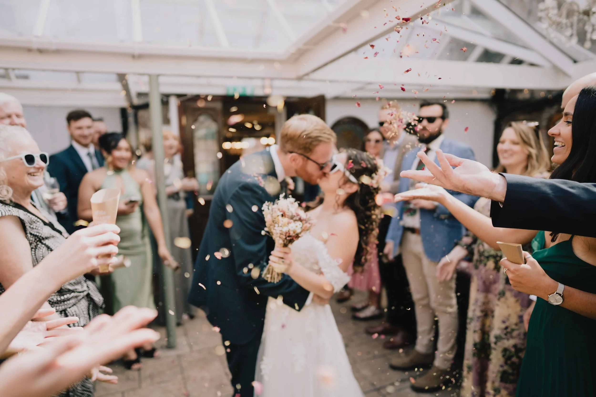 A wedding celebration with a bride and groom kissing, surrounded by guests throwing confetti, indoors with a glass ceiling at Radnor Rooms, Bristol.