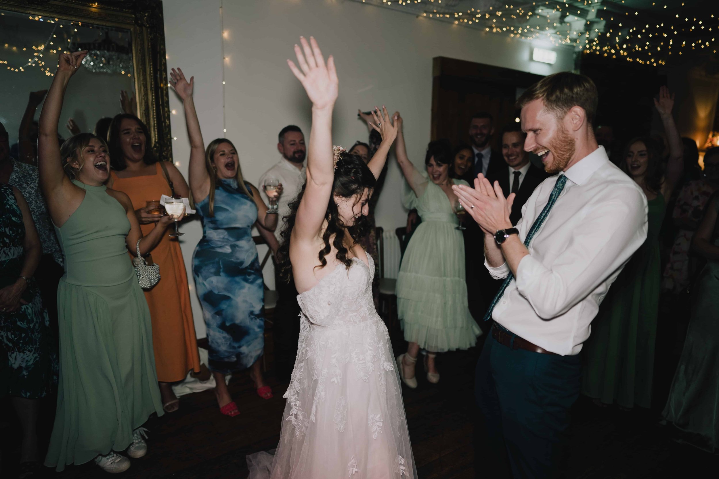 Bride and groom celebrating at their wedding reception, surrounded by friends, with the bride raising her hands and the groom clapping in front of a decorated room with fairy lights at Radnor Rooms, Bristol.