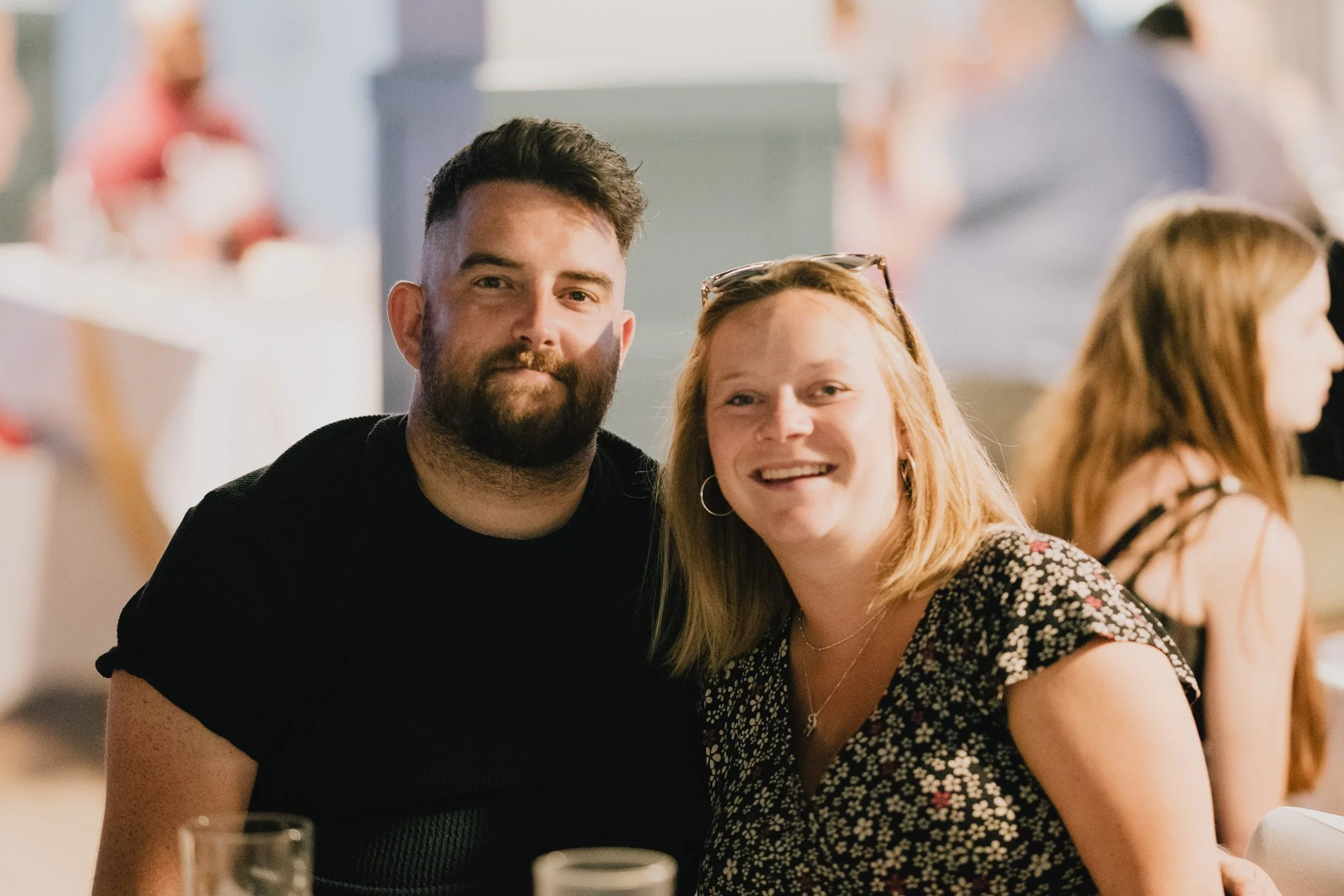 A man and woman smiling at a social event, sitting close together with other people in the background.