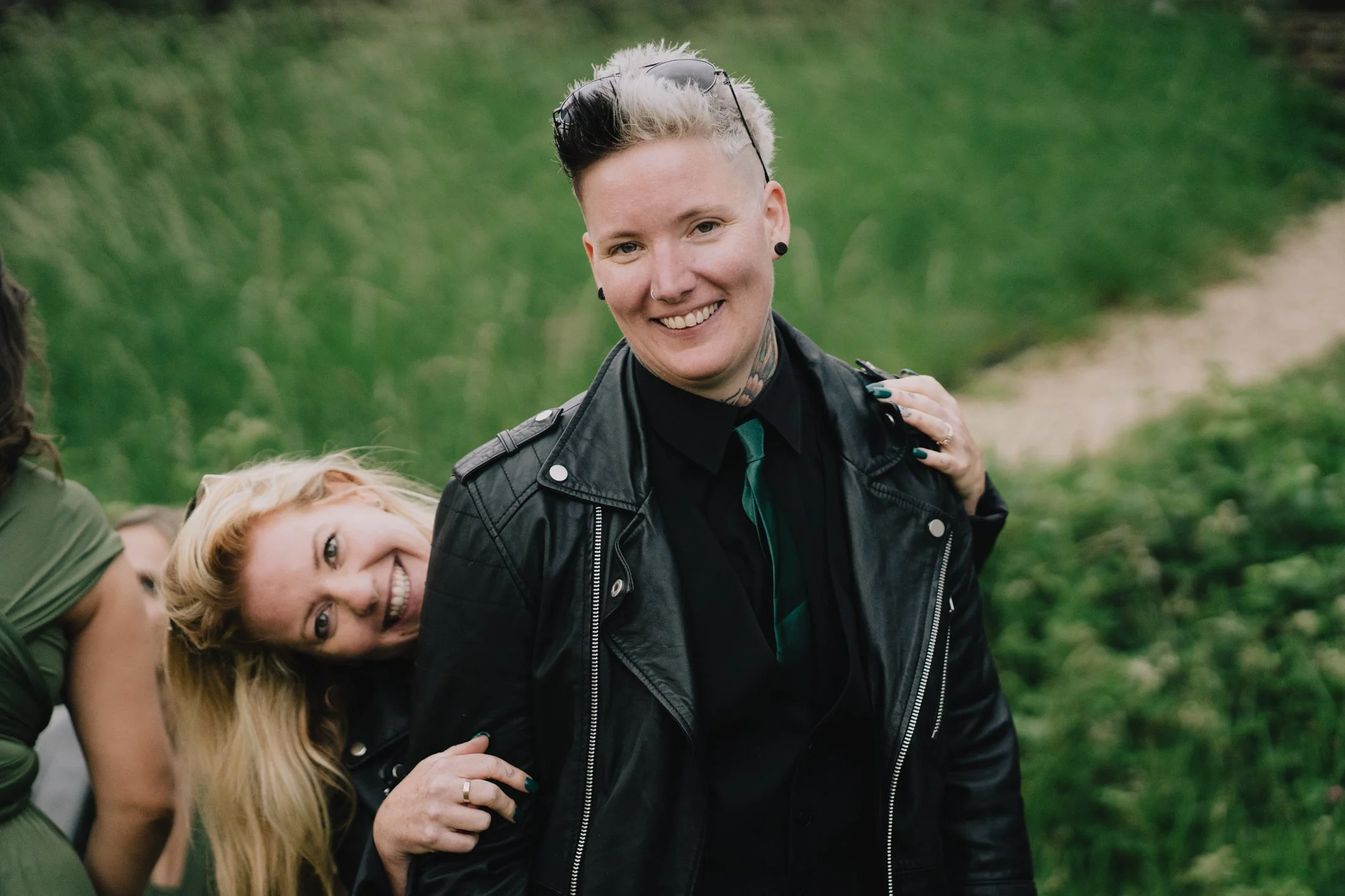 Two smiling women outdoors on a grassy path, with green trees in the background. One woman with short hair and tattoos is in the foreground, wearing a black leather jacket, black shirt, and glasses on her head. The other woman with long blonde hair i