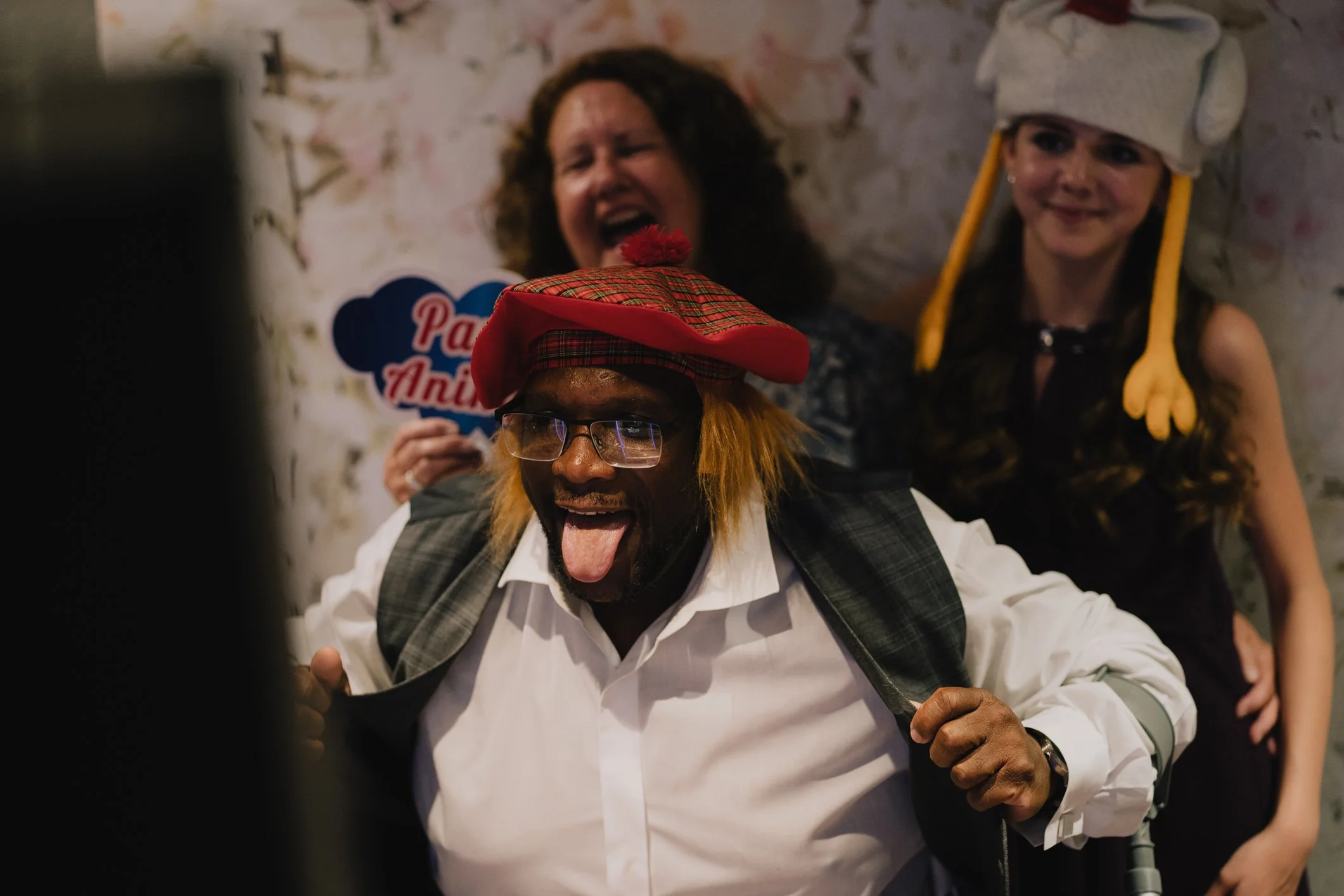 Three people dressed in festive costumes enjoying fun at a celebration. The man in front is sticking out his tongue and wearing glasses, a plaid hat, and a white shirt with a vest. Behind him, a woman with curly hair is laughing and holding a sign th