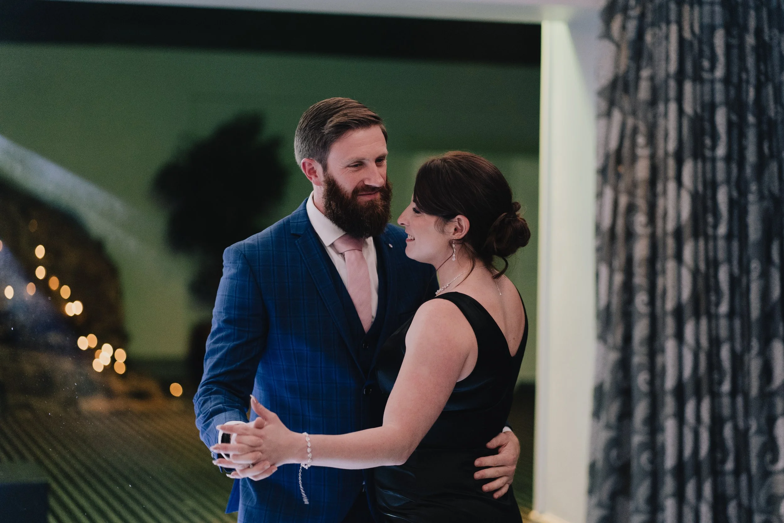 A man and woman dancing closely at a formal event, dressed in elegant attire, with the man wearing a blue plaid suit and the woman in a black dress.