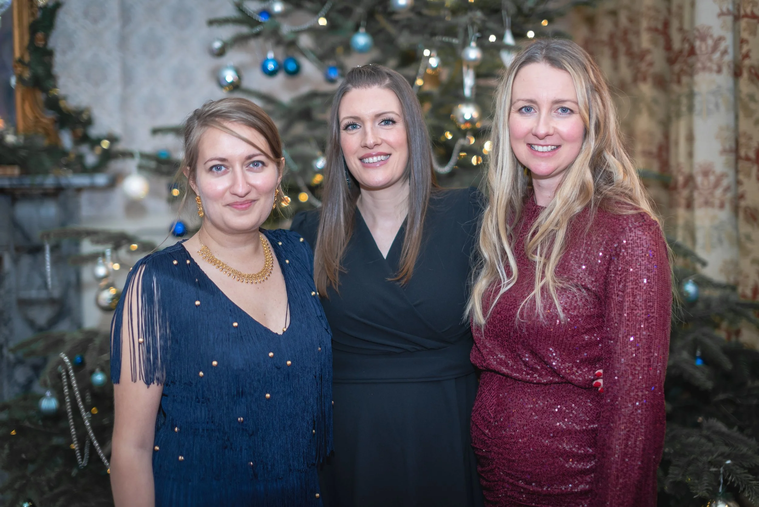 Three women in festive dresses standing in front of a decorated Christmas tree.