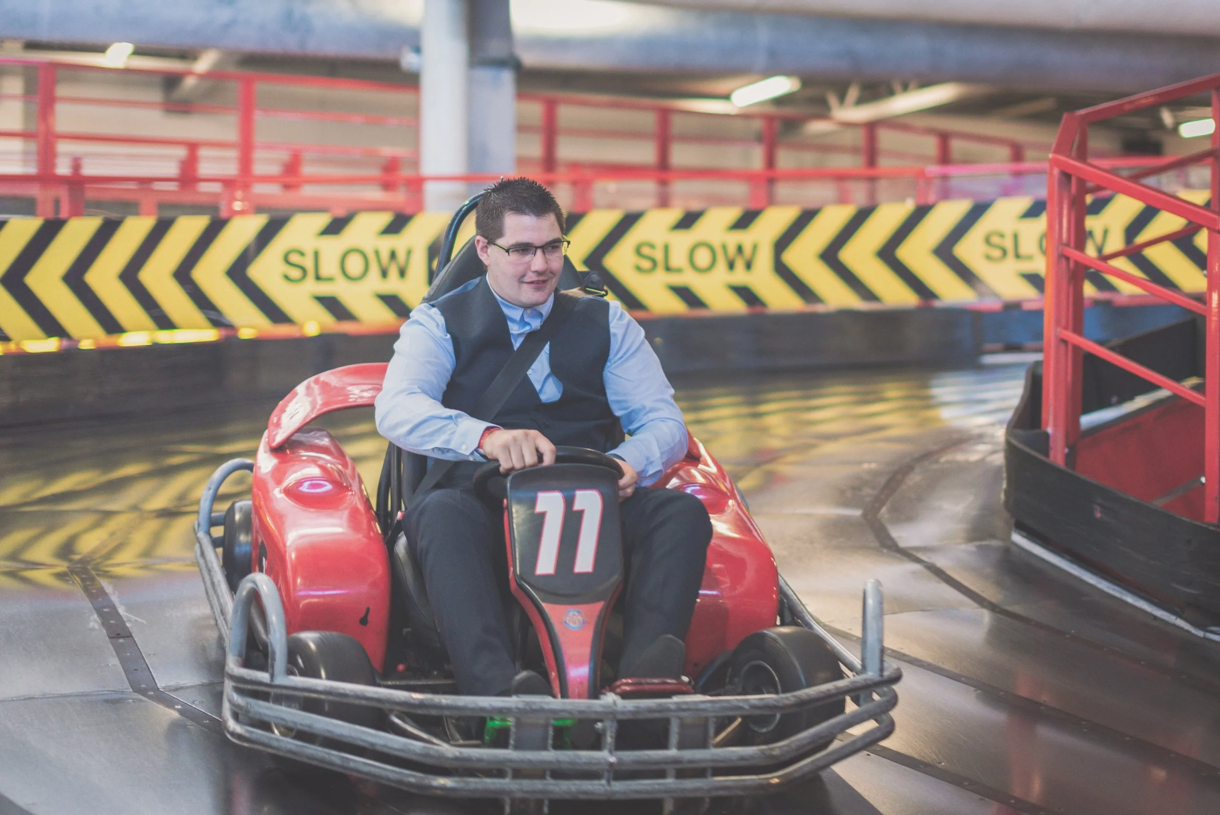 A man in business attire driving a small red go-kart inside an indoor racing facility.