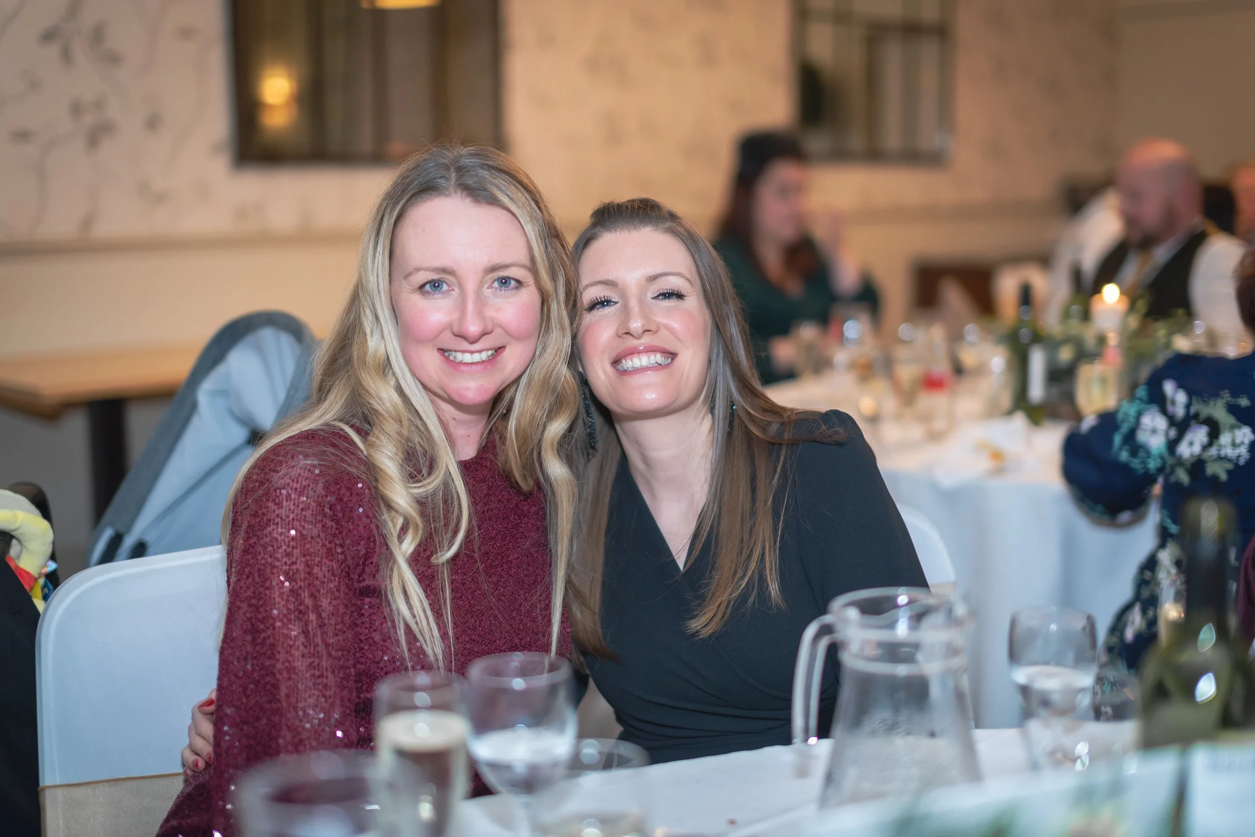 Two women smiling and sitting together at a dinner table during an indoor event or celebration.