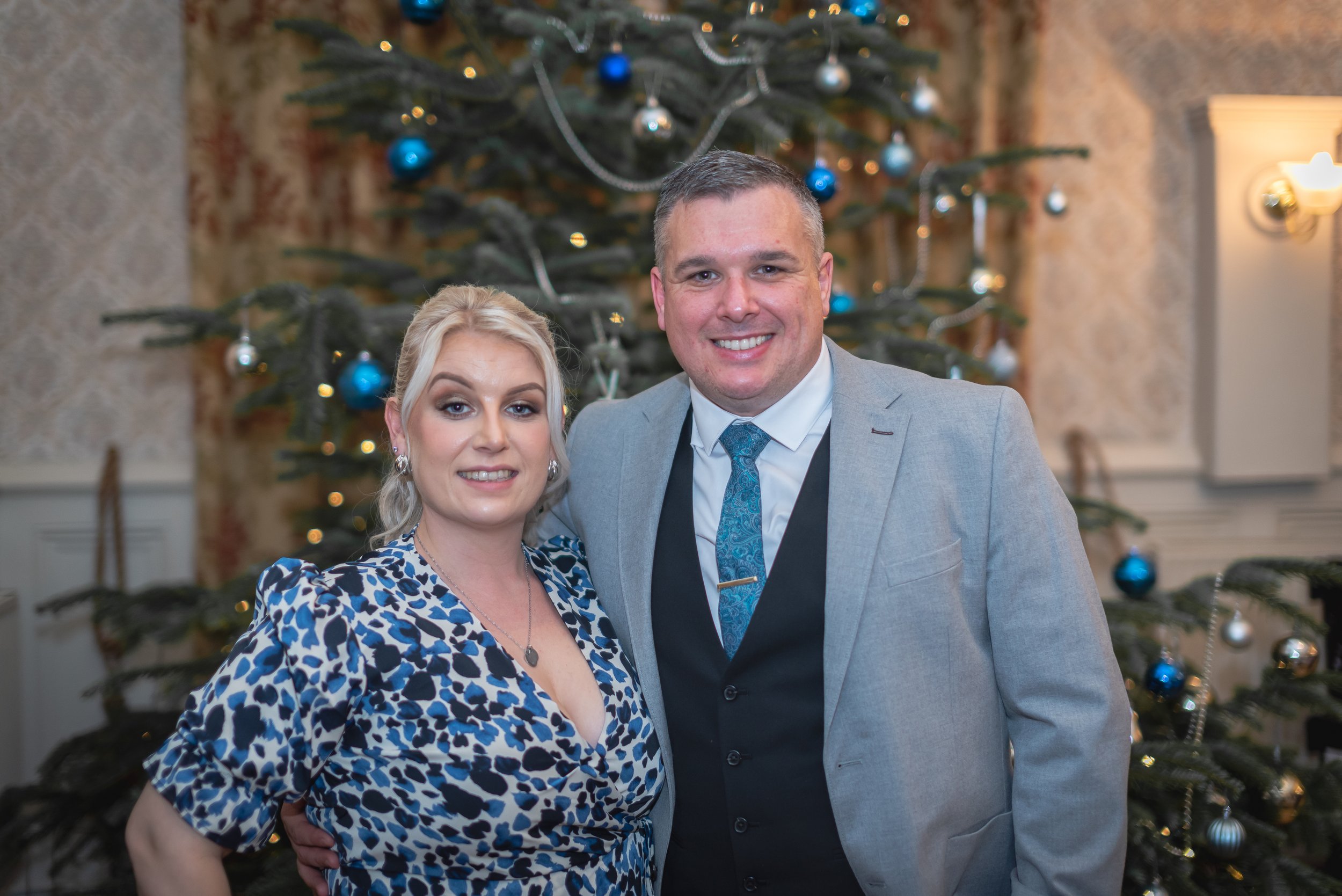 A smiling man and woman standing close together in front of a decorated Christmas tree, dressed in festive attire for a holiday celebration.