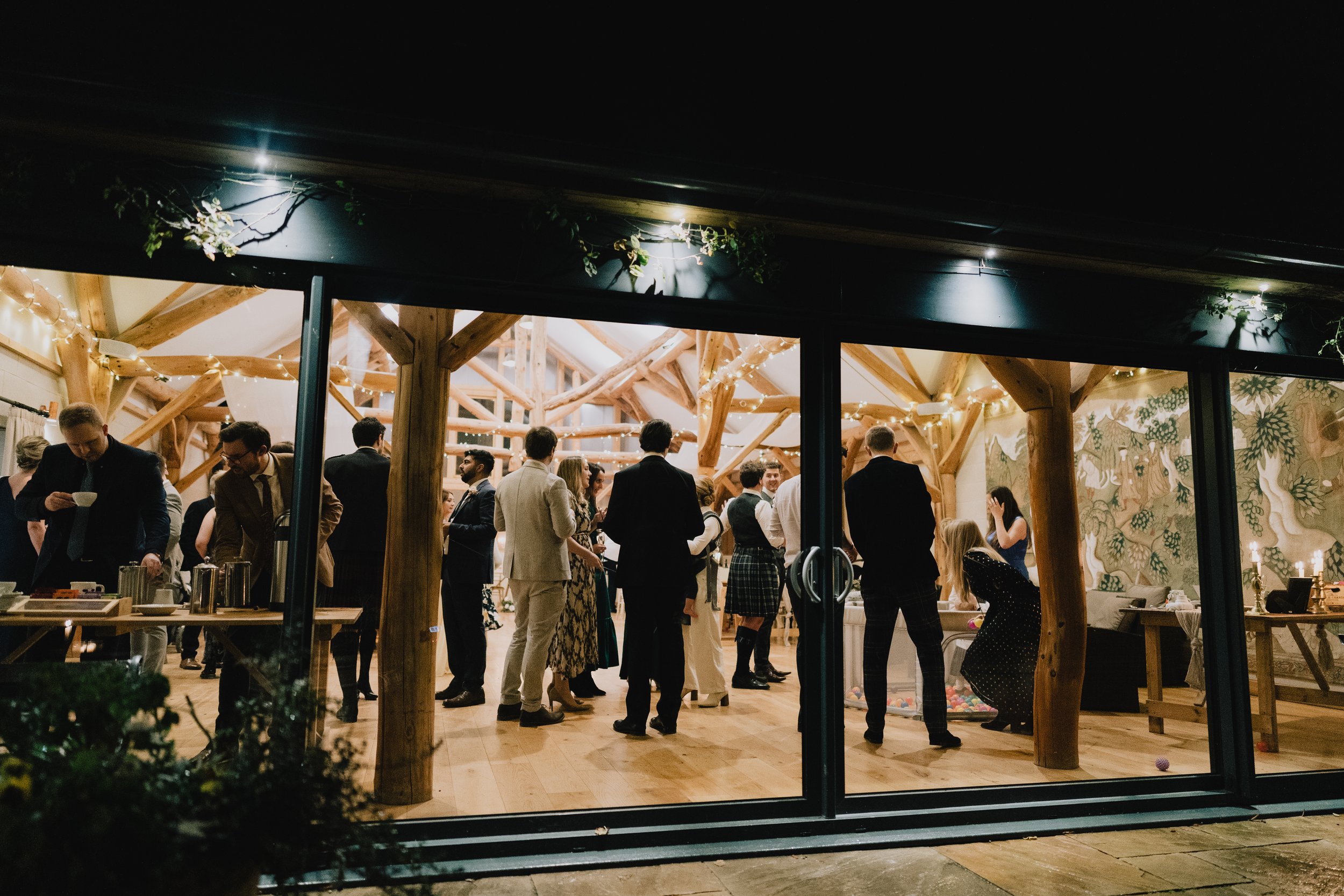 People gathering indoors at a wooden event space with string lights, some dancing, some chatting, and a woman playing with balloons.