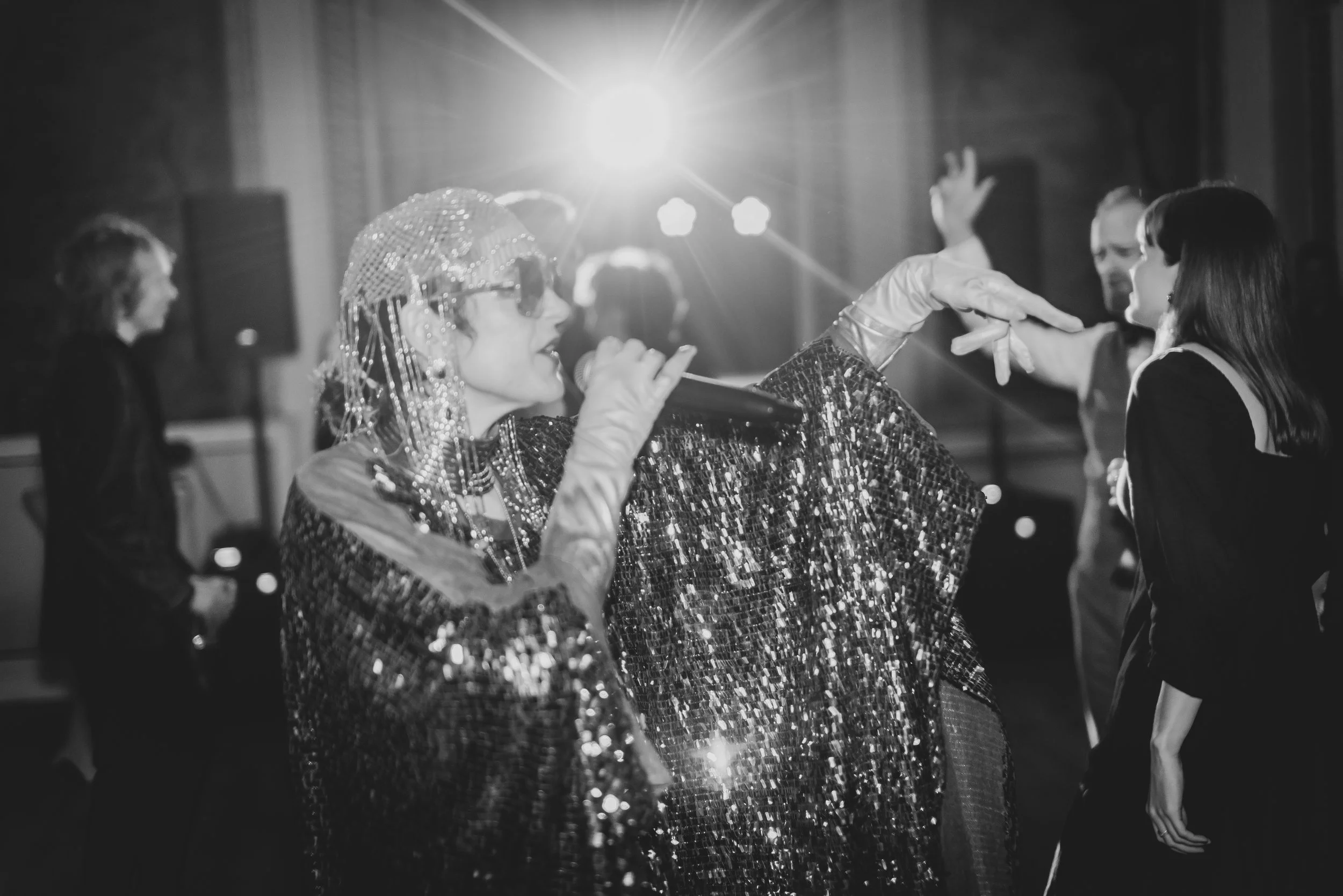 A woman dressed in retro style with sequined clothing, wearing sunglasses and a shiny headpiece, singing into a microphone at a party or event. She is surrounded by people dancing and socializing, with a bright light source behind her creating a star